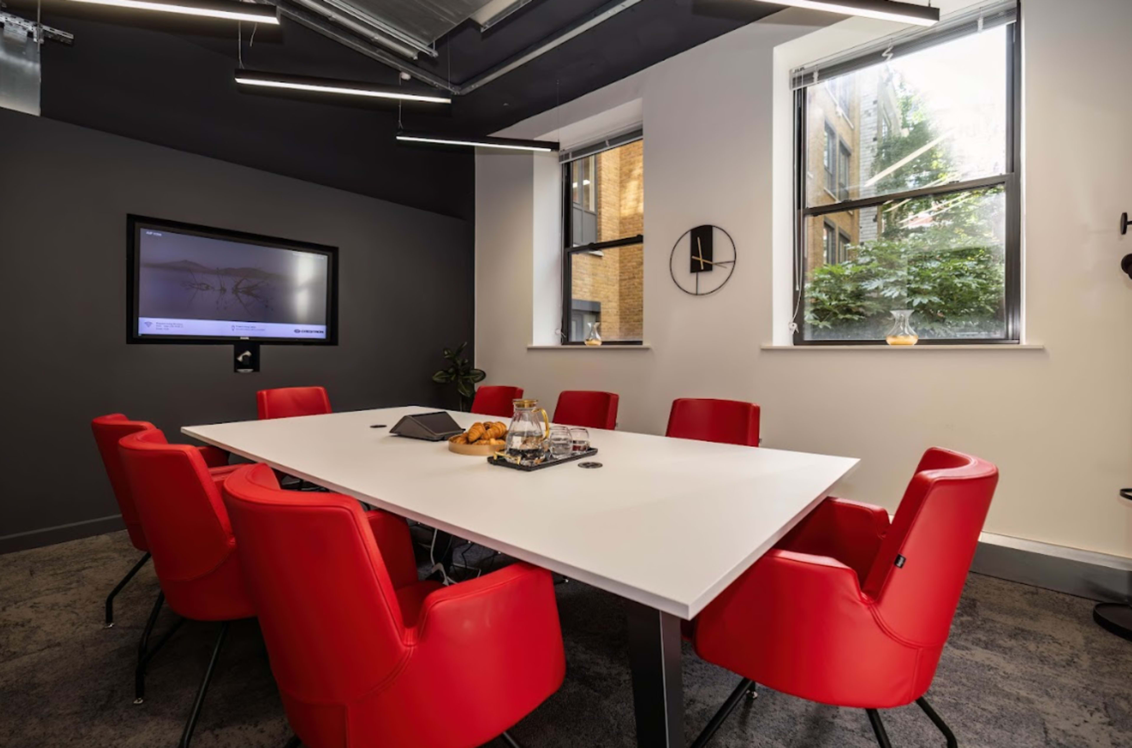 A modern conference room features a white table surrounded by six red chairs, with a large screen on the wall and windows allowing natural light.