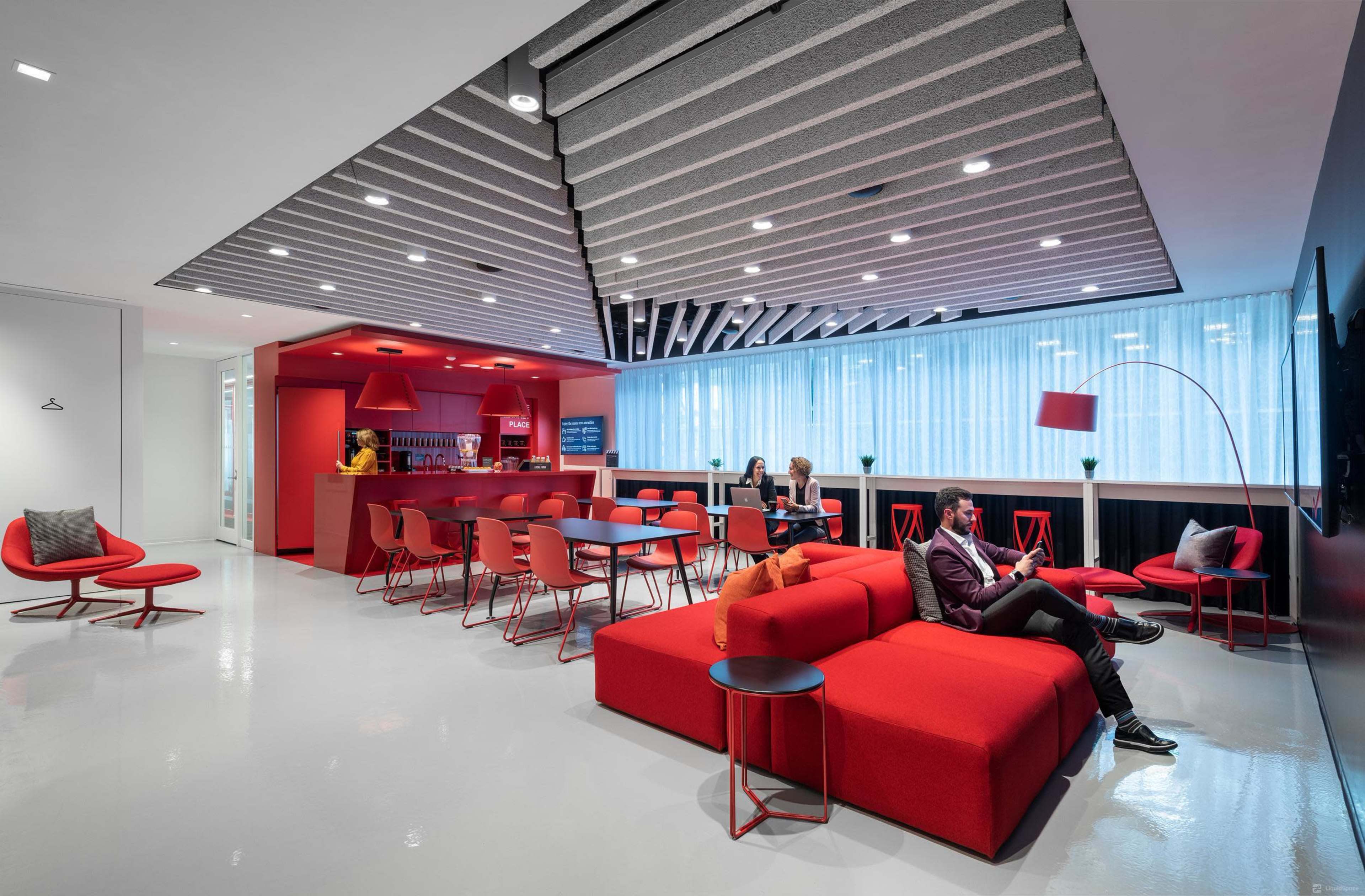 A modern lounge area features red furniture, a kitchen bar, and people engaged in conversation and reading.