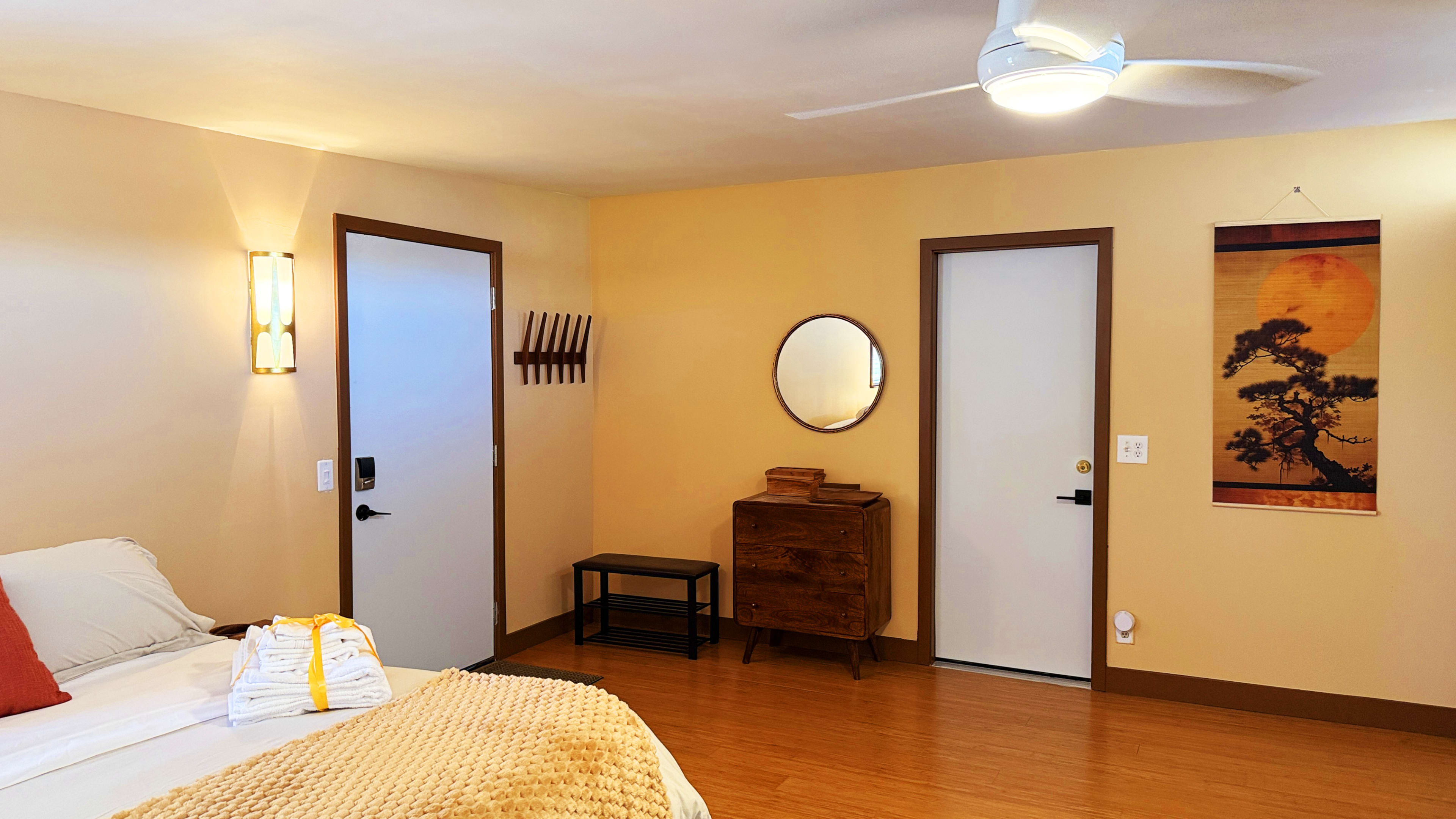 The image shows a simple bedroom with a bed, a wooden dresser, a round mirror on the wall, and a door with a coat rack nearby.