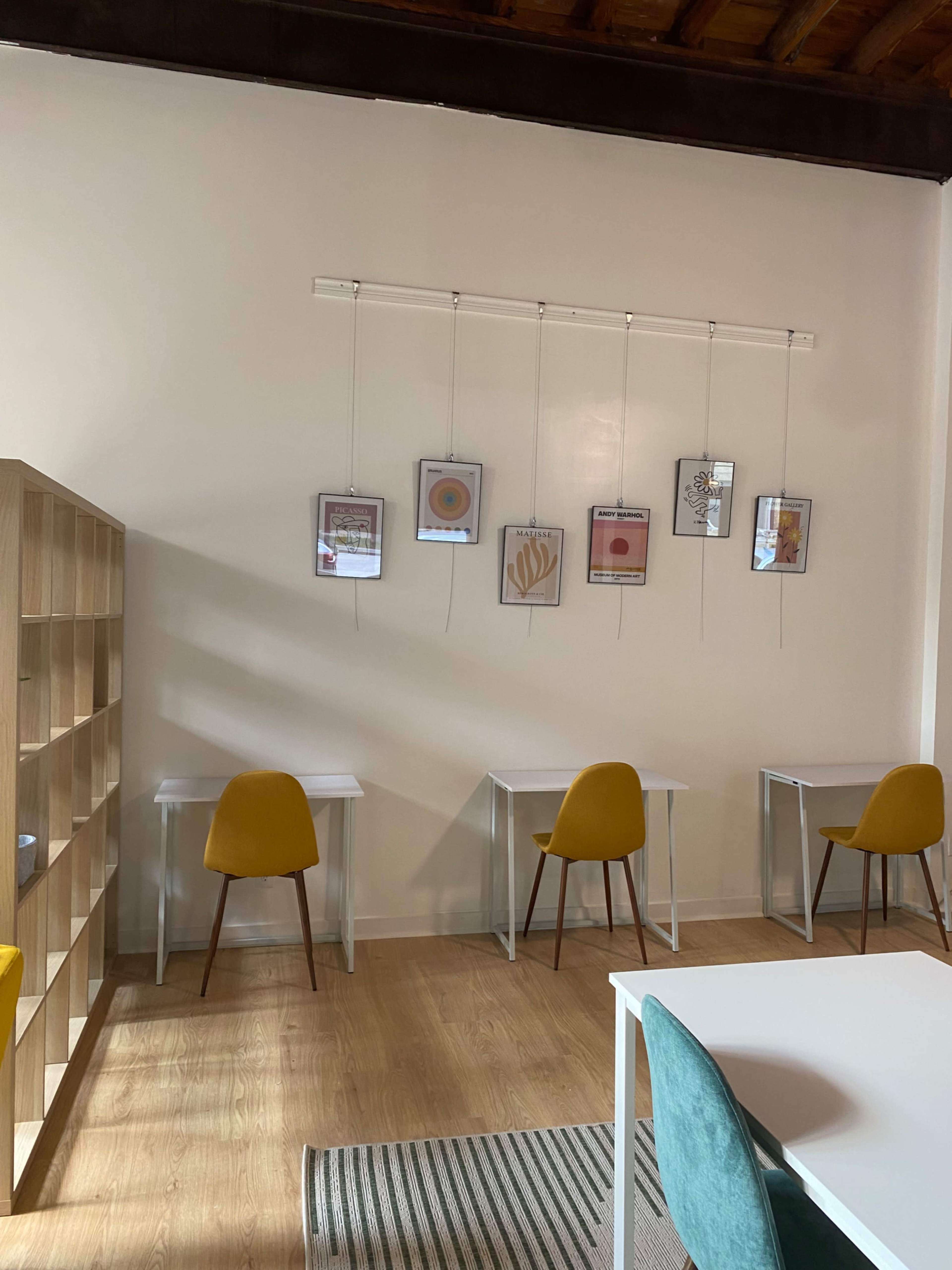 A minimalist workspace with four yellow chairs arranged around white tables, accompanied by framed artworks hanging on the wall above.