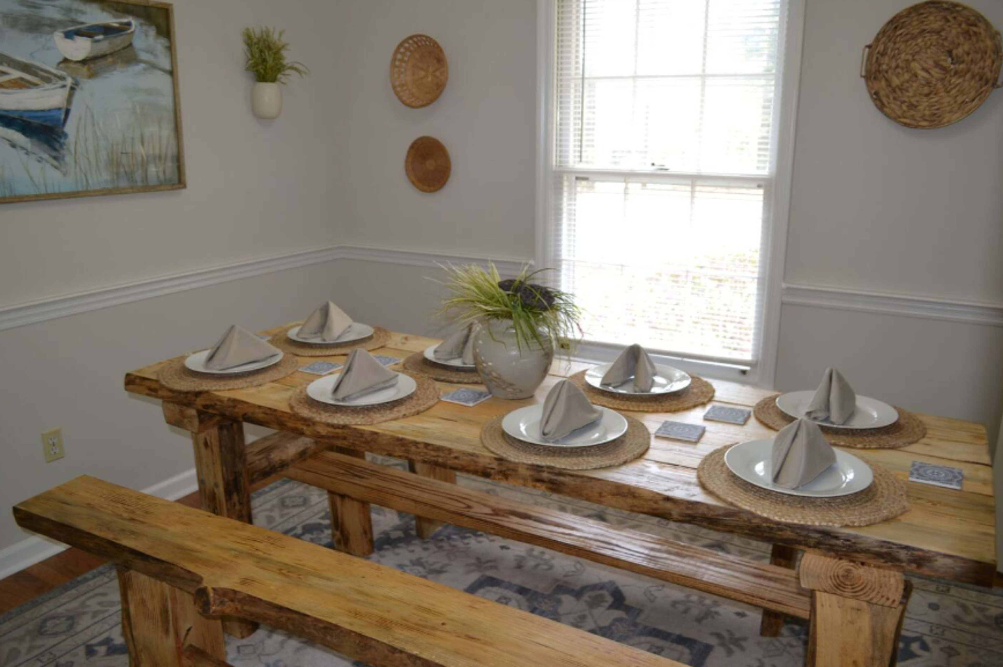 A rustic wooden dining table is set with plates and neatly folded napkins, surrounded by a bench and chairs, with a vase of greenery as a centerpiece.