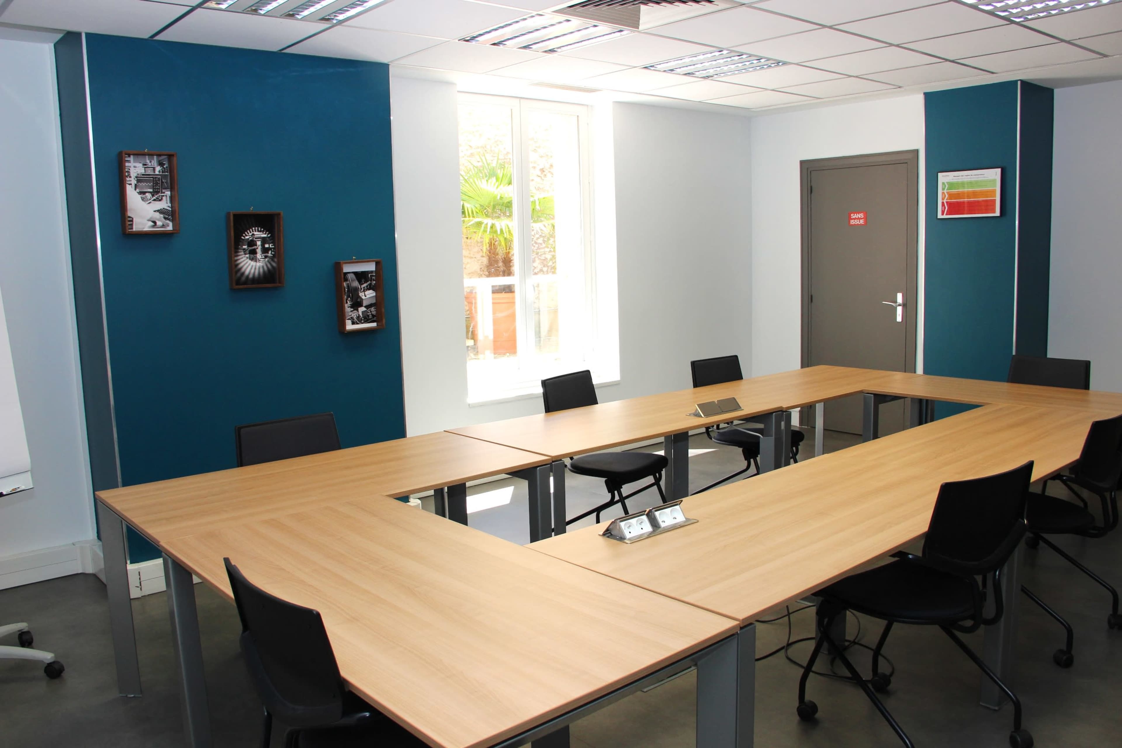 A rectangular conference room with a large wooden table surrounded by black chairs, featuring blue and gray walls and a window letting in natural light.
