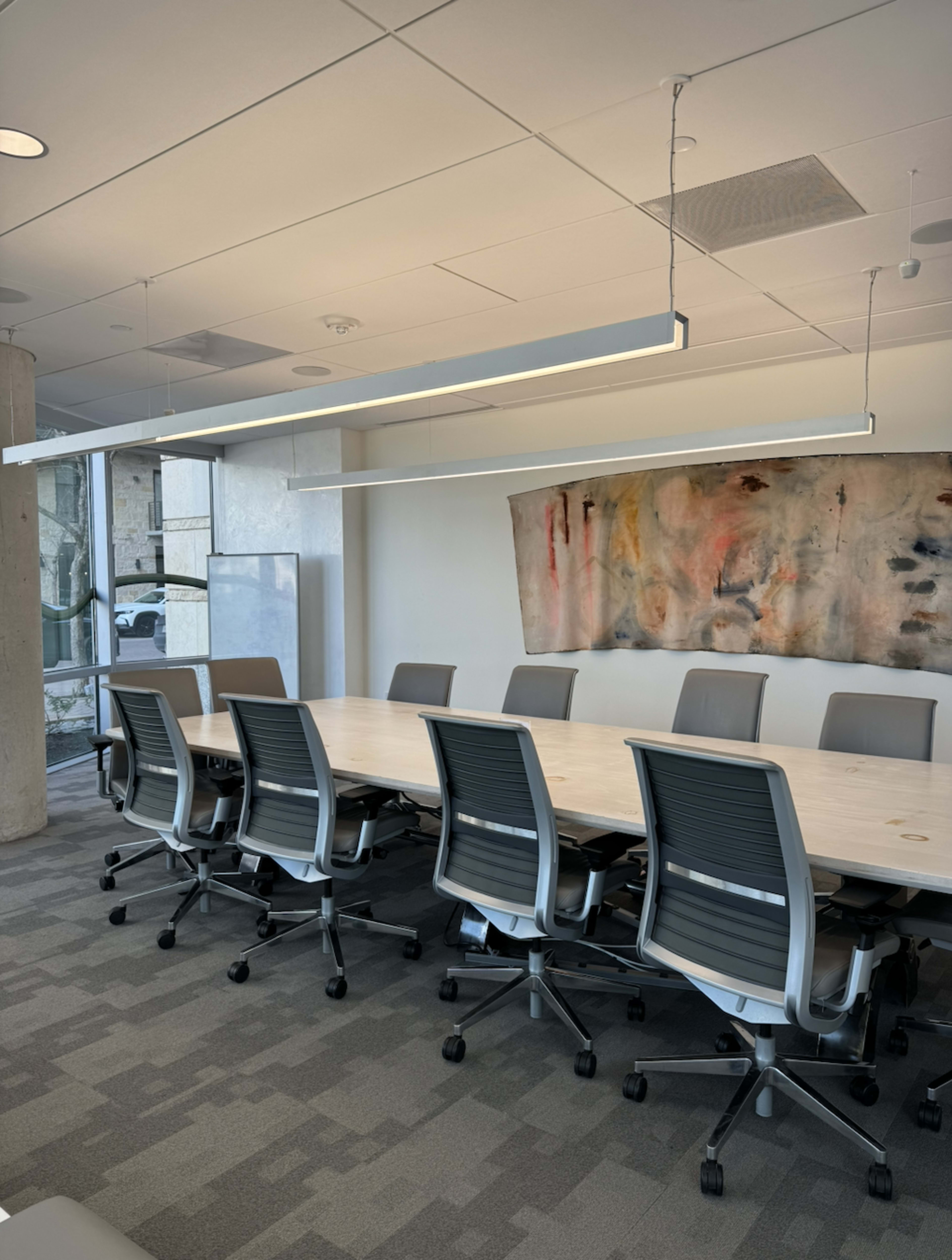 A modern conference room features a long wooden table surrounded by ergonomic chairs, with overhead lighting and a large abstract painting on the wall.