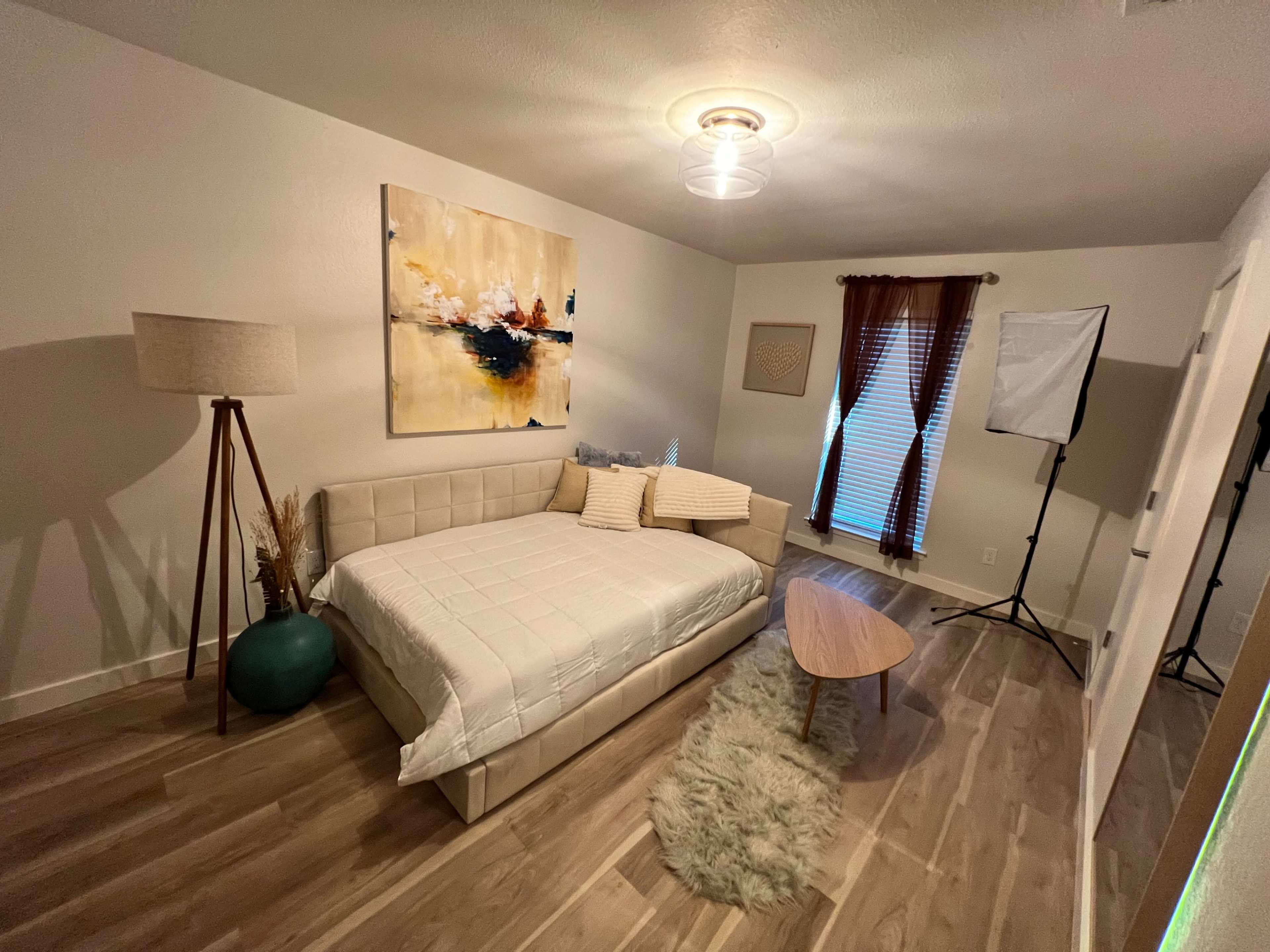 A neatly arranged bedroom featuring a light-colored sofa bed, a wooden coffee table, a modern lamp, and decorative art on the wall, with natural light streaming through the window.