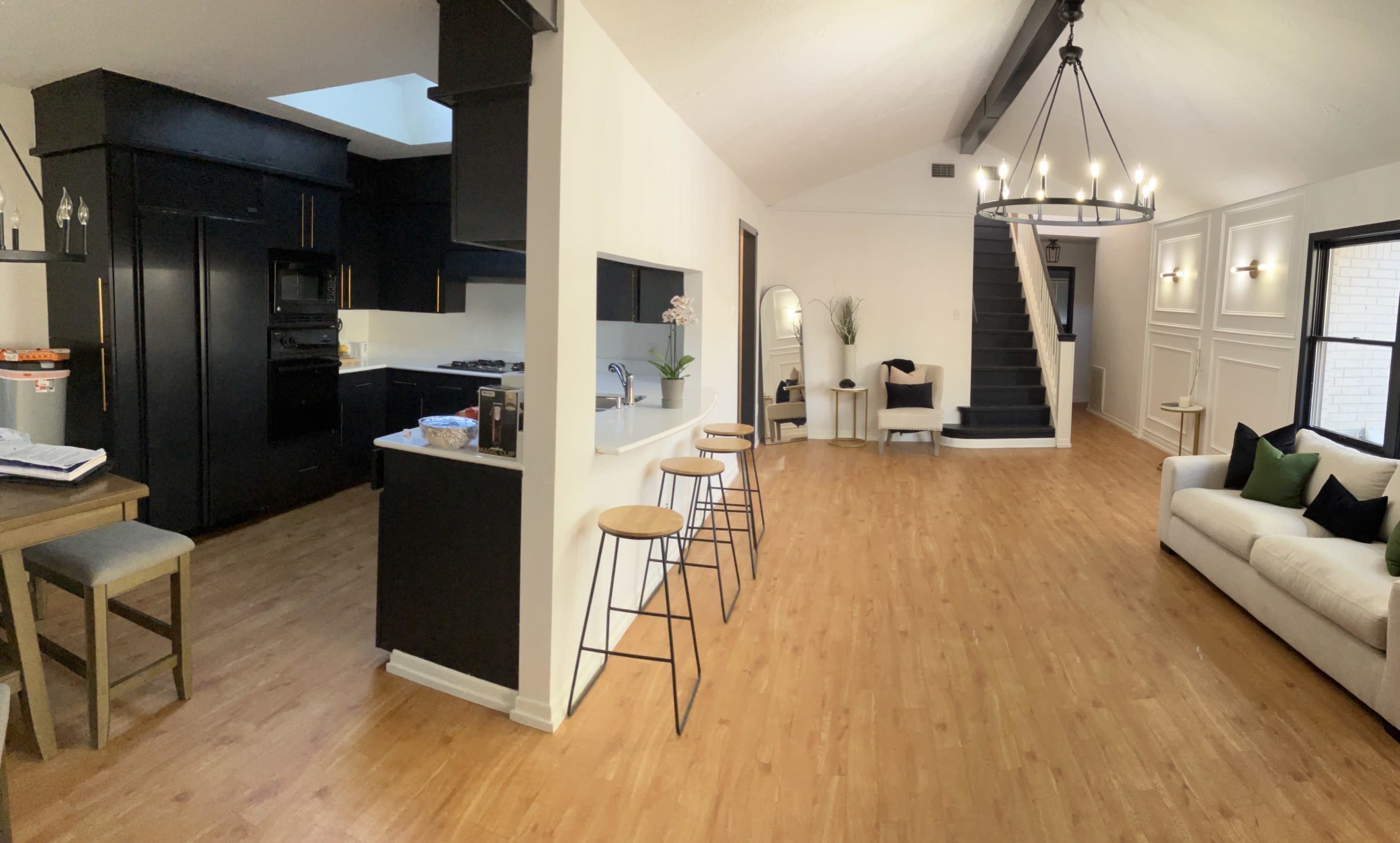 A modern open-concept living space featuring a kitchen with black cabinetry, a bar with three stools, and a staircase leading to an upper level.