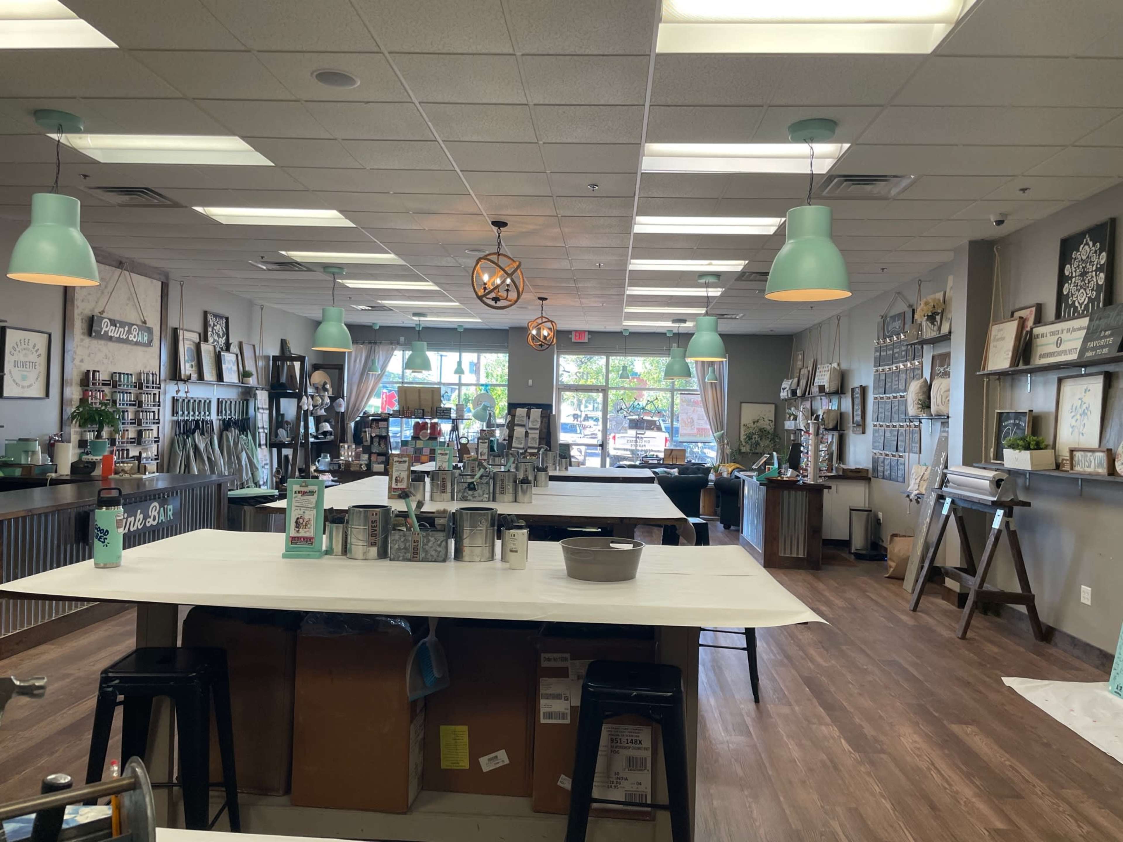 The image shows a spacious, well-lit craft studio with tables, various supplies, and decorative wall art, featuring large windows that allow natural light to enter.