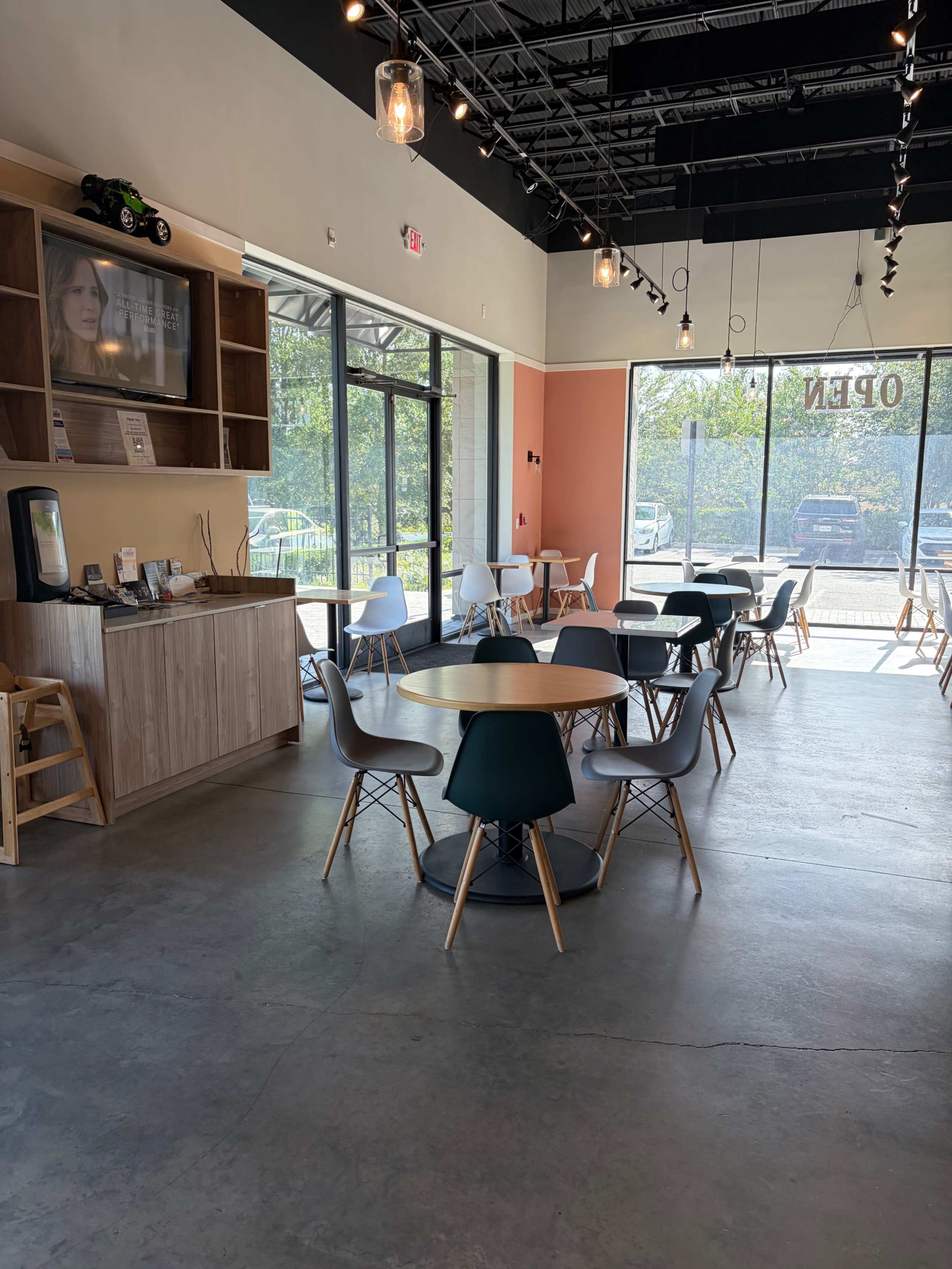 The image shows a spacious café interior with tables and chairs arranged, large windows allowing natural light, and a service area with a display.