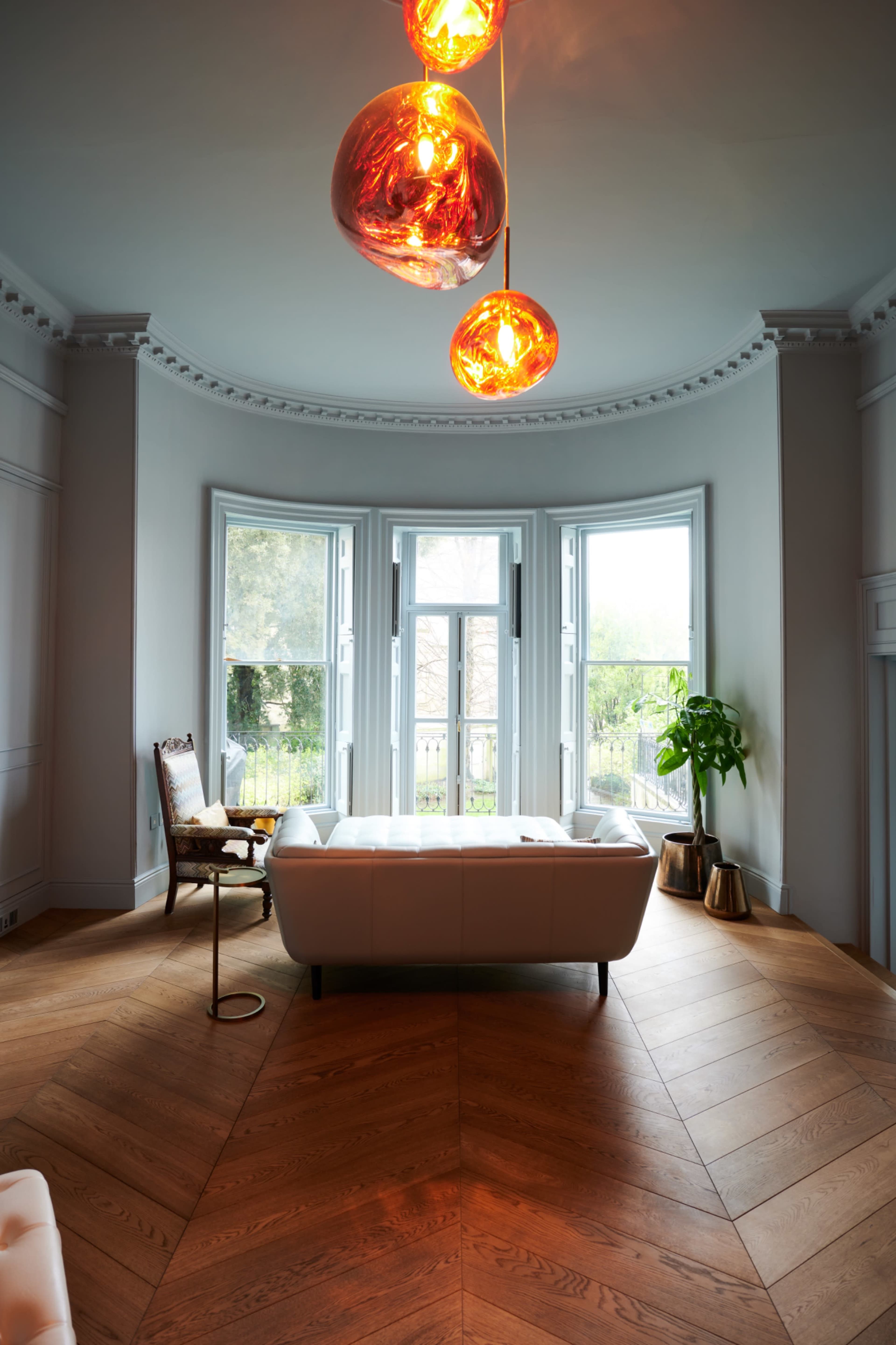 A modern living room features a white sofa, a wooden chair, large windows allowing natural light, and pendant lights above a herringbone-patterned wooden floor.
