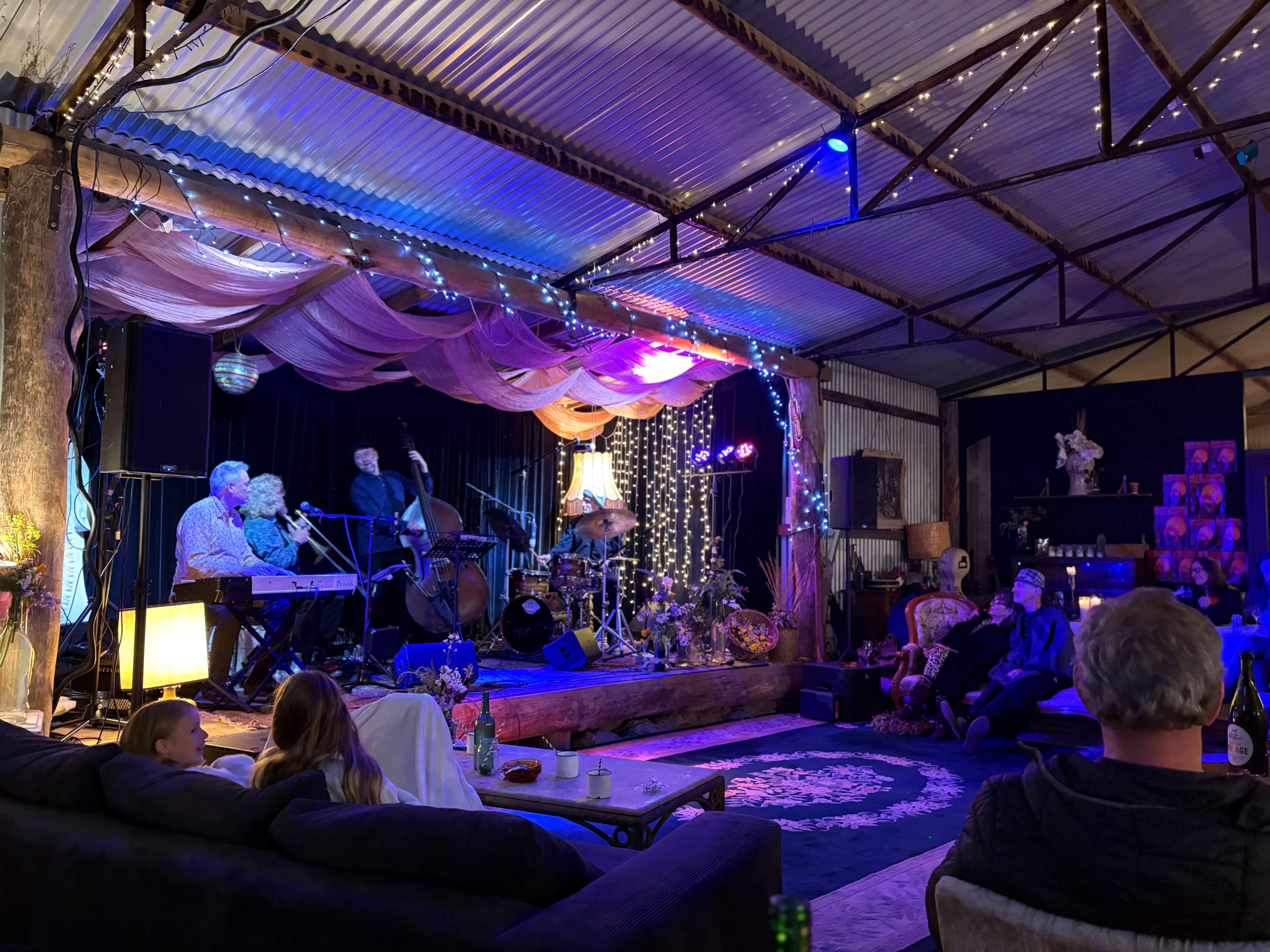 A live music performance is taking place in a dimly lit venue, featuring a small band with a piano, bass, and drums, while an audience enjoys the show from scattered seating.