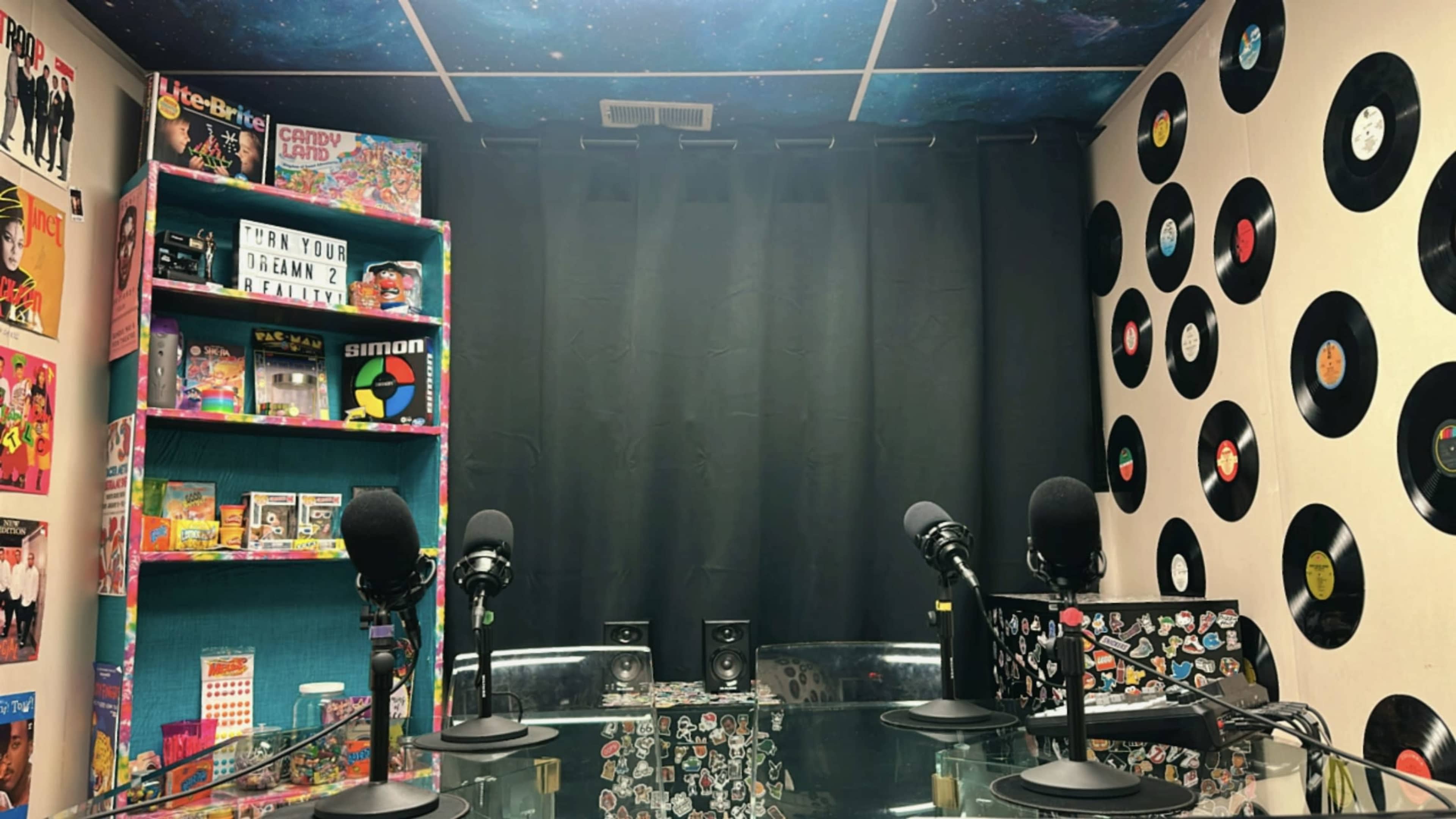 The image shows a podcast recording studio featuring colorful shelves filled with vintage games and toys, a black curtain backdrop, and several microphones arranged around a glass table.