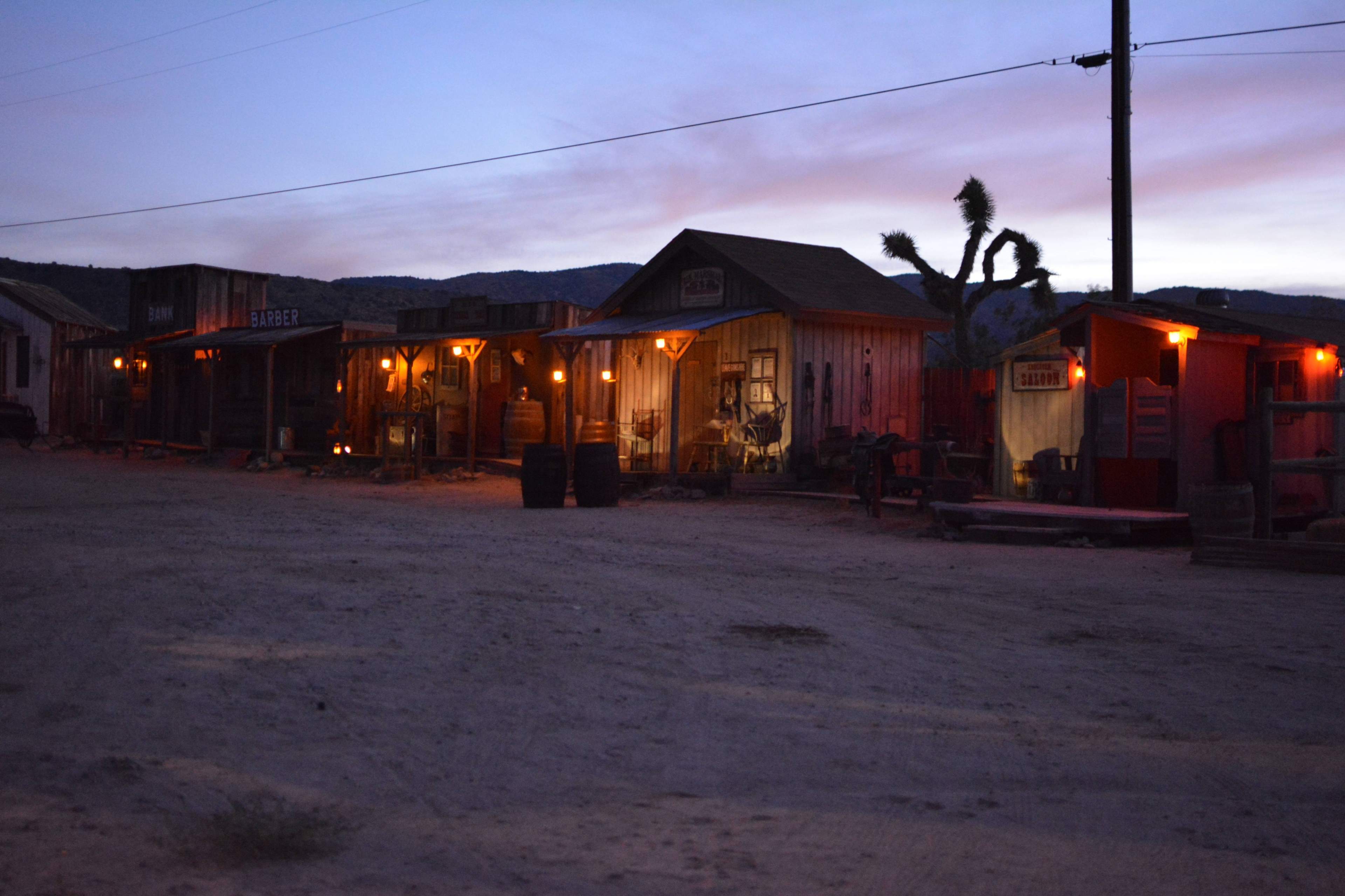 The scene depicts a western-style ghost town at dusk, with illuminated storefronts and a dirt pathway under a colorful sky.