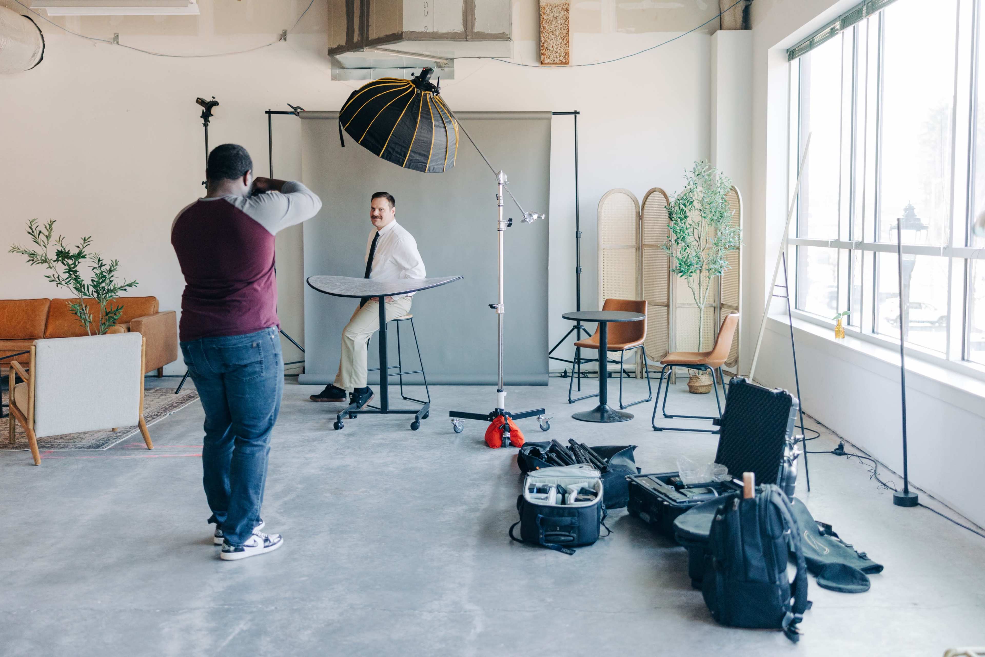 A photographer captures a portrait of a man sitting on a stool in a well-lit studio with various photography equipment and props arranged around the space.