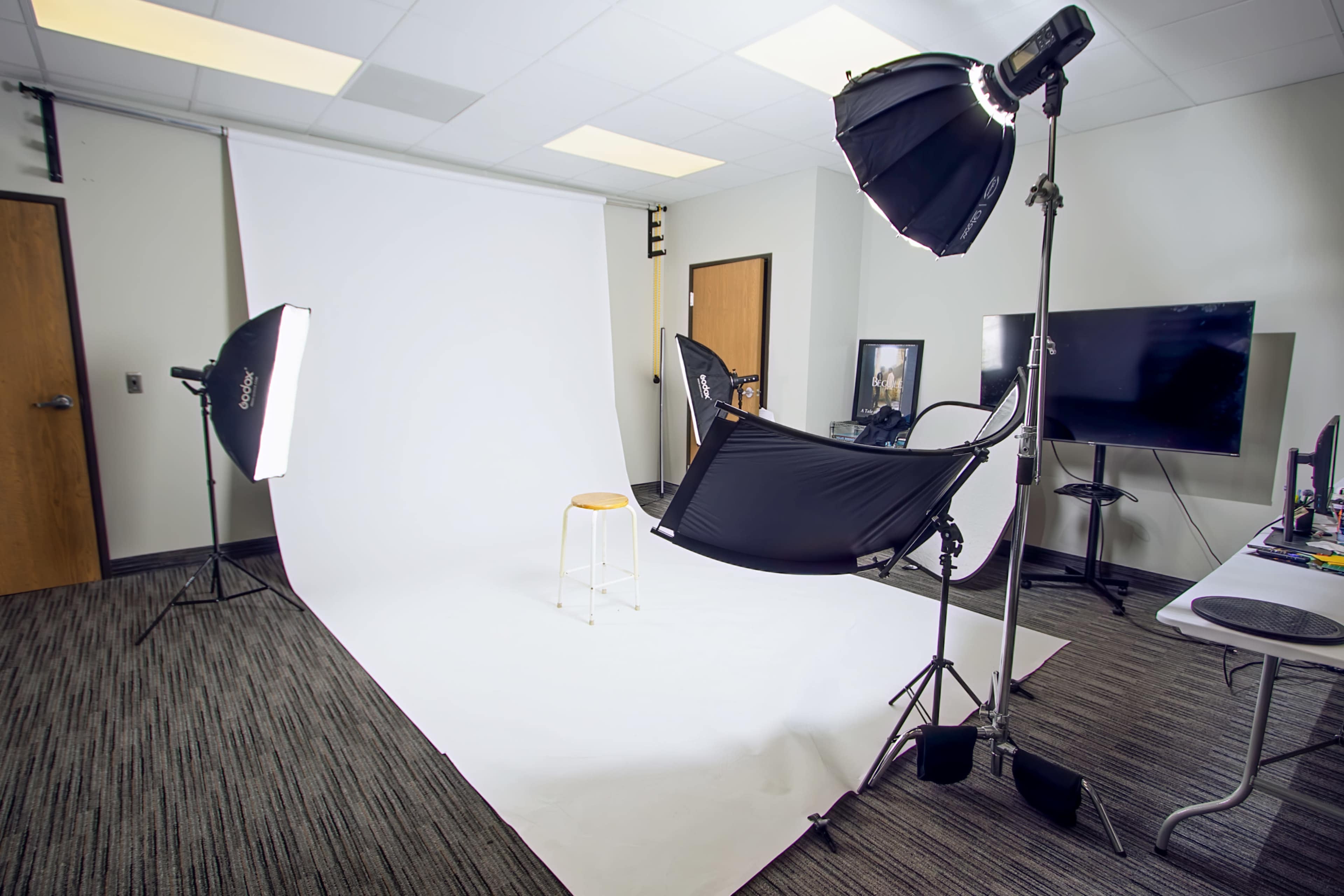 The image shows a photography studio setup with a white backdrop, a stool, and softbox lighting equipment.