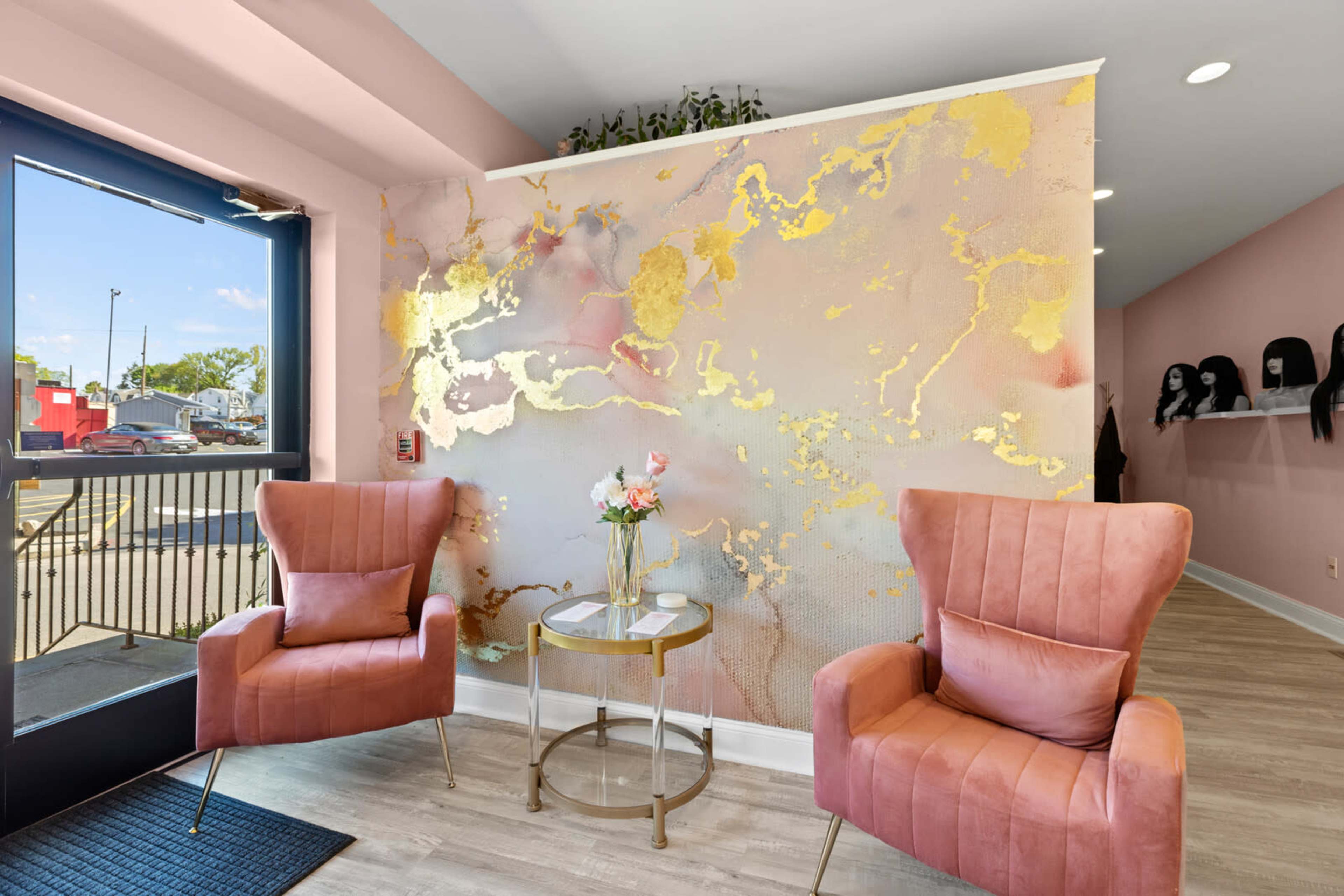 A modern salon interior with pink walls, two velvet armchairs, a gold accent table, and a decorative mural.