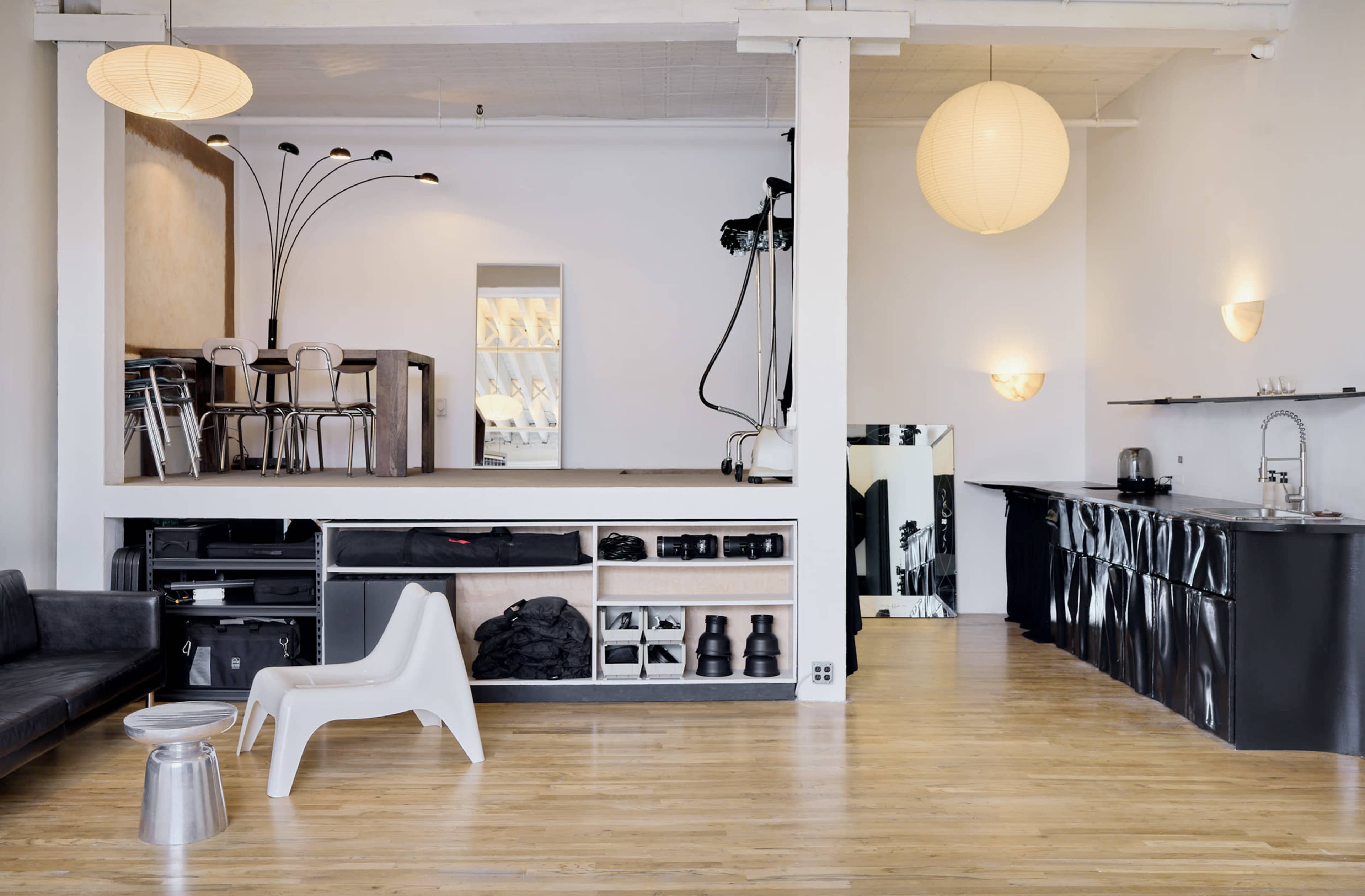 The image shows a modern, open-plan living space featuring a kitchen area, a sitting area with a black couch, and a dining section with tall chairs and a light fixture.
