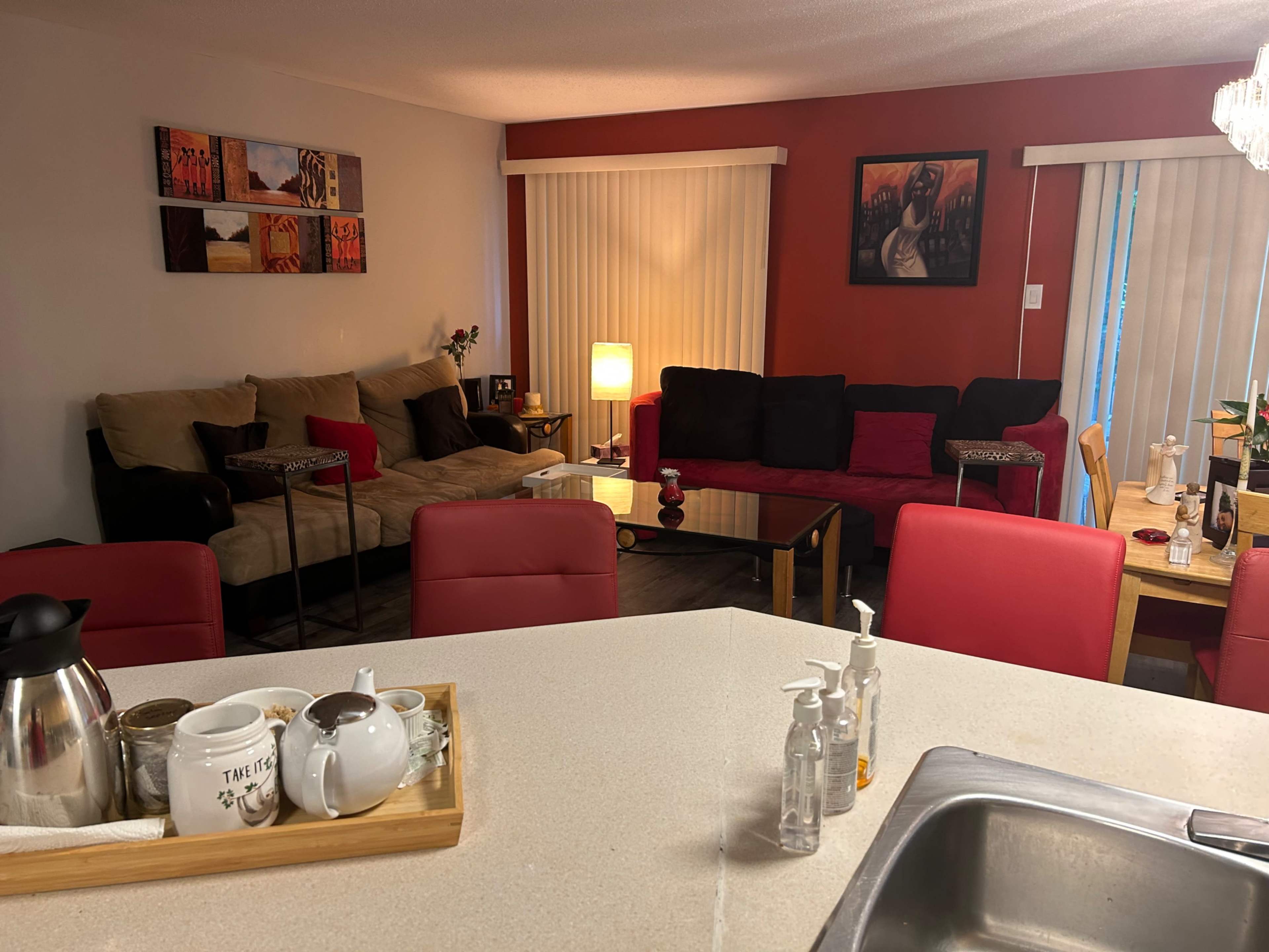 The image shows a living space with a beige couch and a red sectional, a table in the center, and a kitchen area visible in the foreground with various utensils.