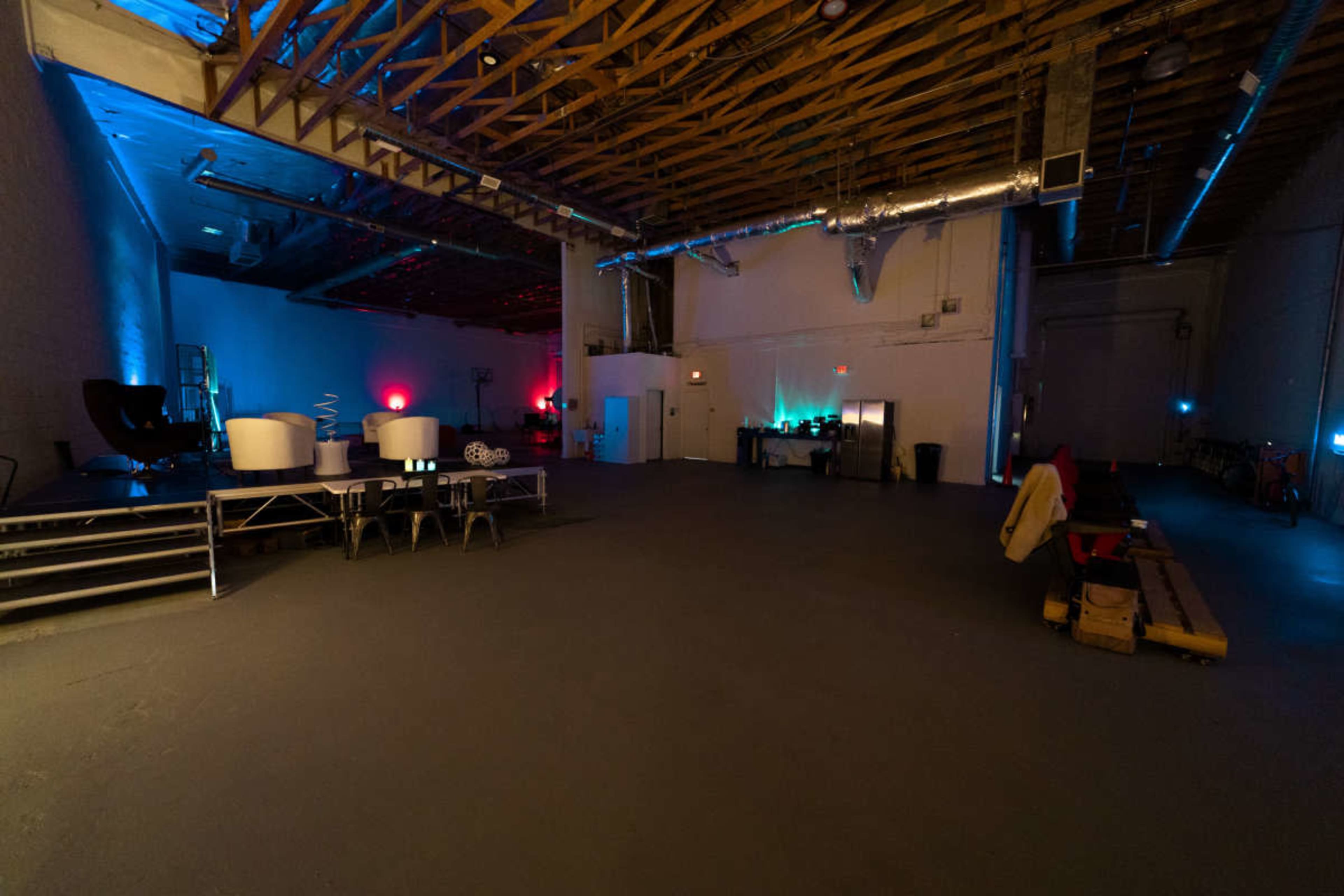 The image shows a spacious room with a high ceiling, illuminated by colored lights, featuring empty seating areas and tables at one end.