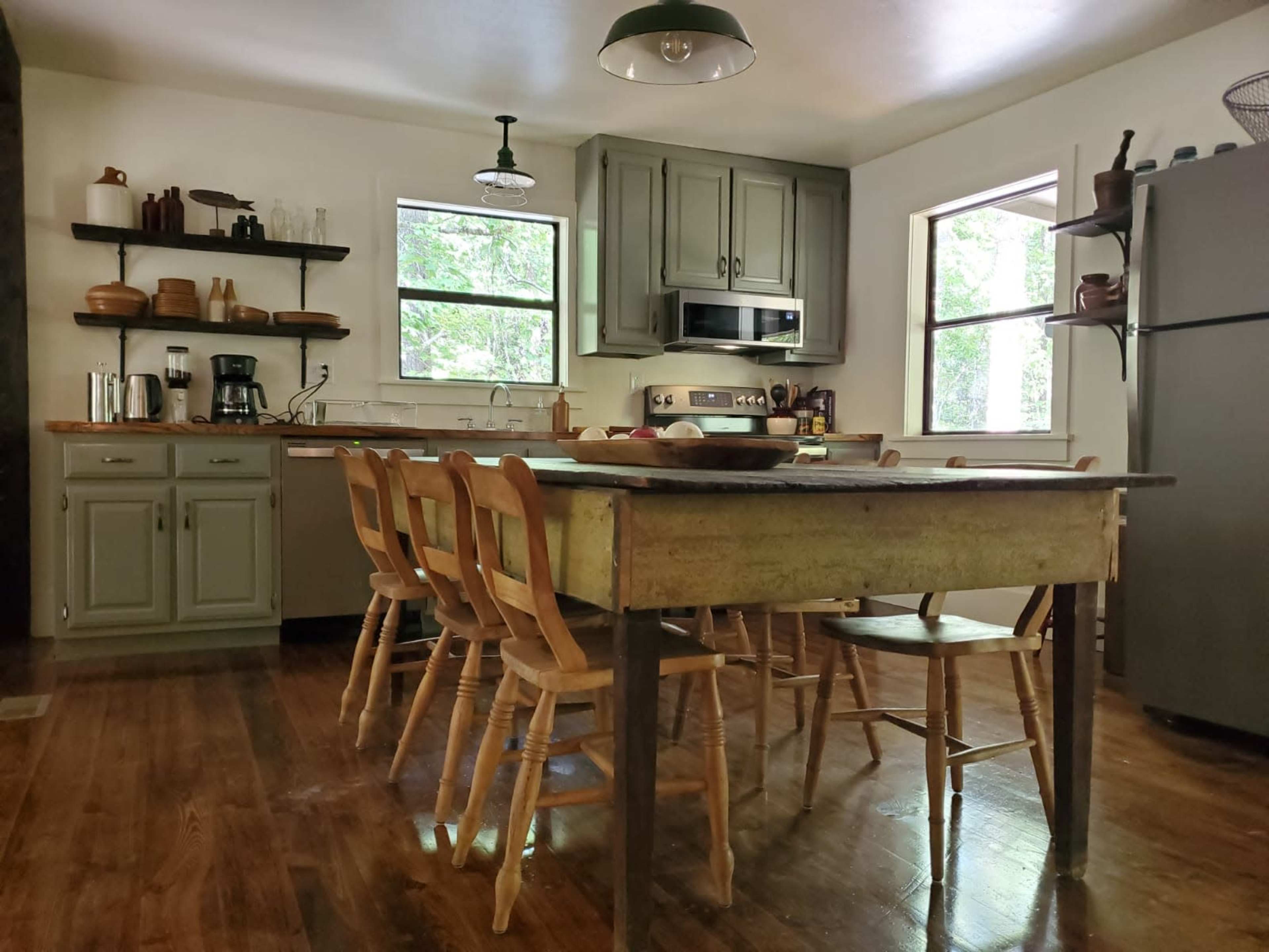 The kitchen features a wooden table surrounded by six chairs, with cabinets, a stove, and shelves displaying dishes against a backdrop of large windows.