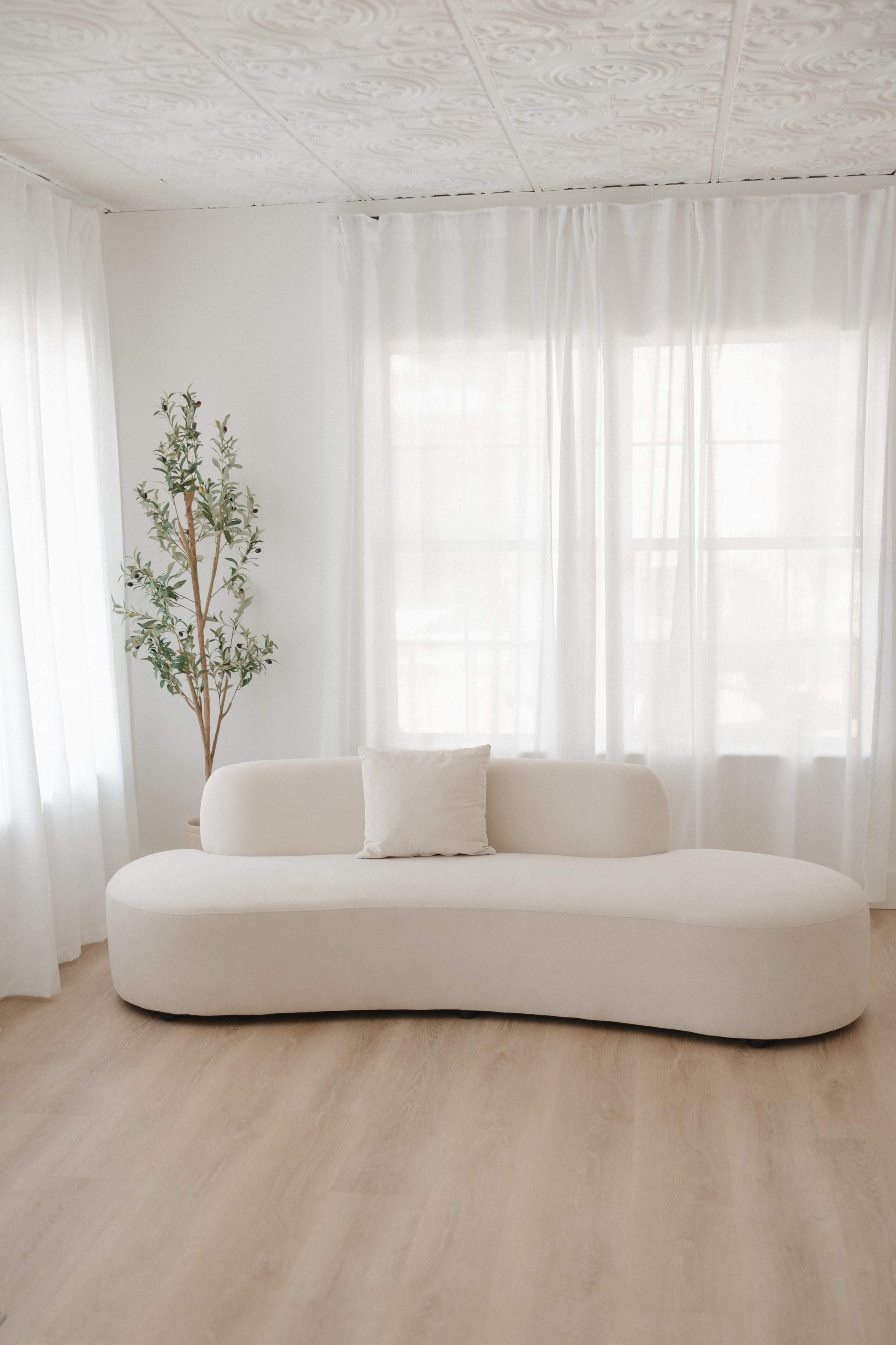 A curved, cream-colored sofa is positioned in a bright room with large windows and sheer white curtains, complemented by a small indoor plant.