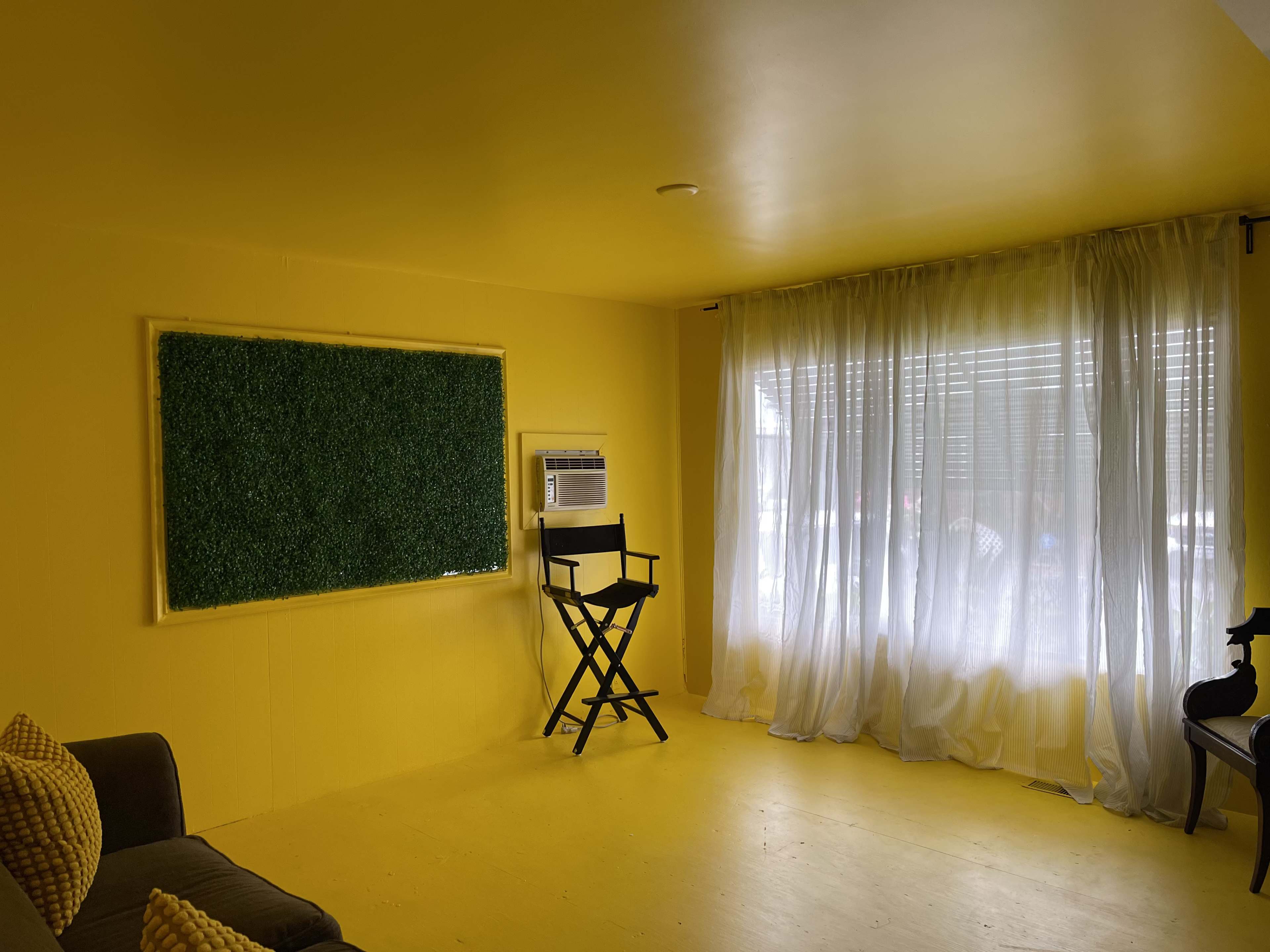 The room features yellow walls, a green wall decoration, a window with sheer curtains, and a director's chair positioned near the window.