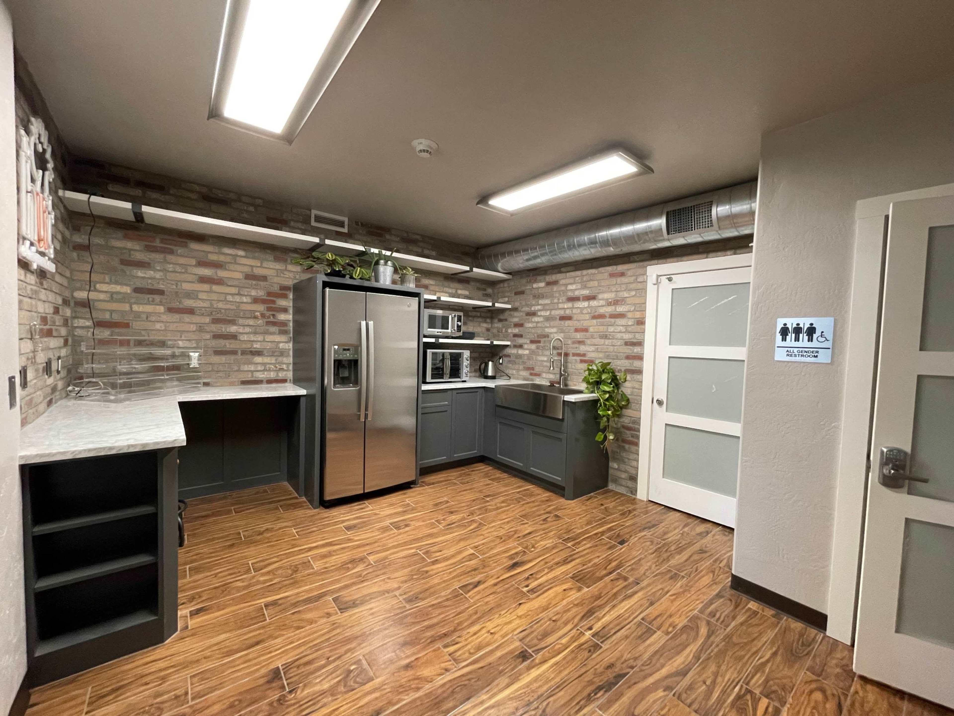 The image shows a modern kitchen setup with stainless steel appliances, including a refrigerator and microwave, set against a brick wall and wood-patterned flooring.