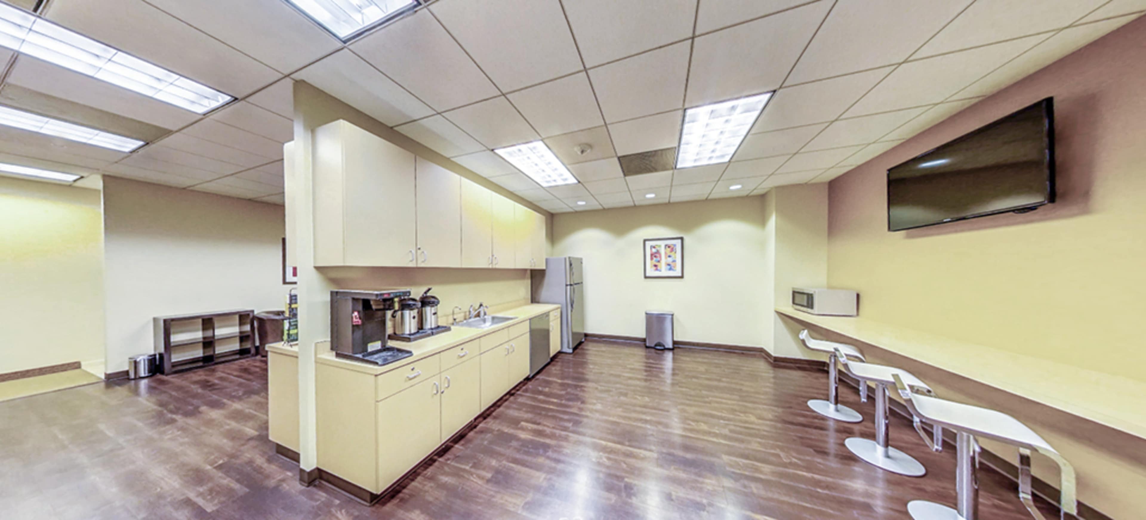 A modern office kitchenette with a coffee station, refrigerator, and stools along a counter, all under bright overhead lighting.
