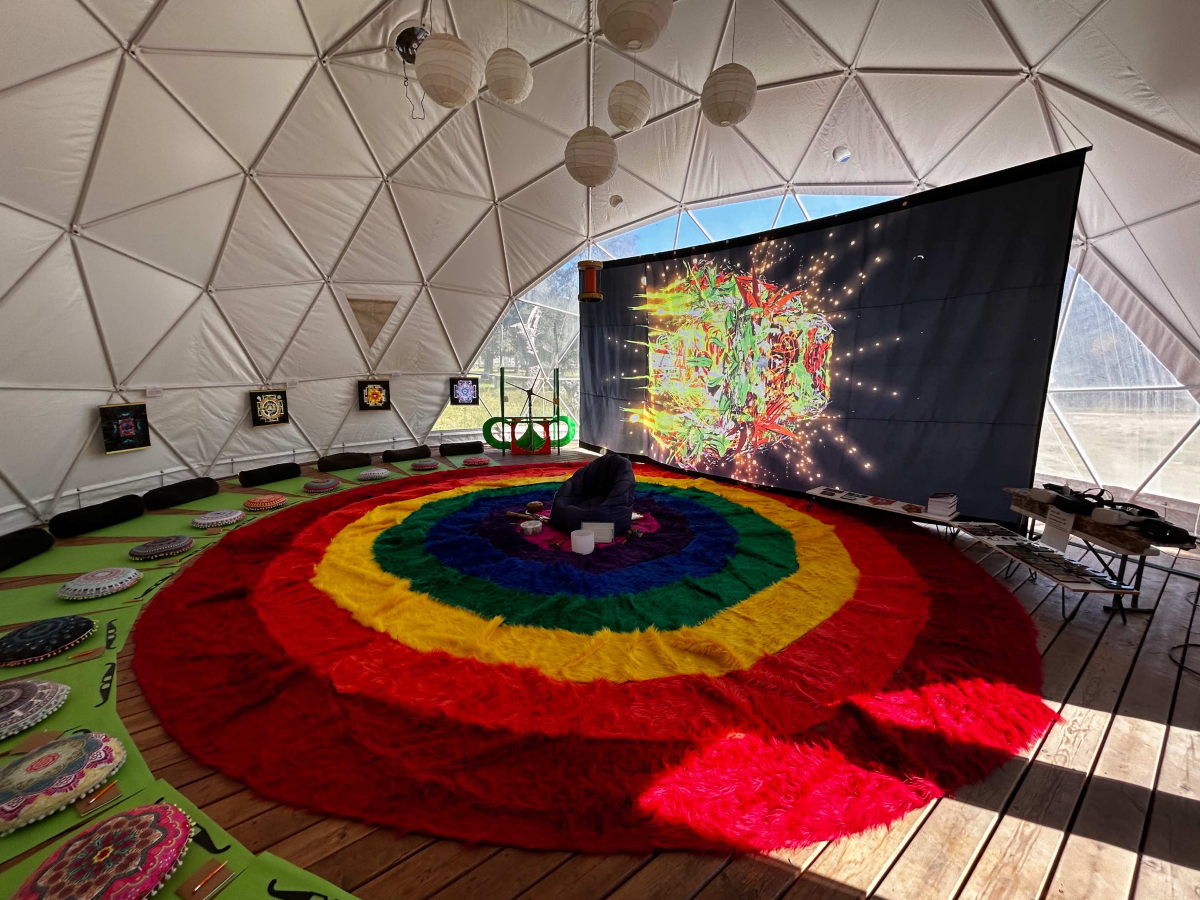 The image shows the interior of a geodesic dome featuring a large circular colorful rug at the center, surrounded by seating and a backdrop with vibrant artwork displayed on a screen.
