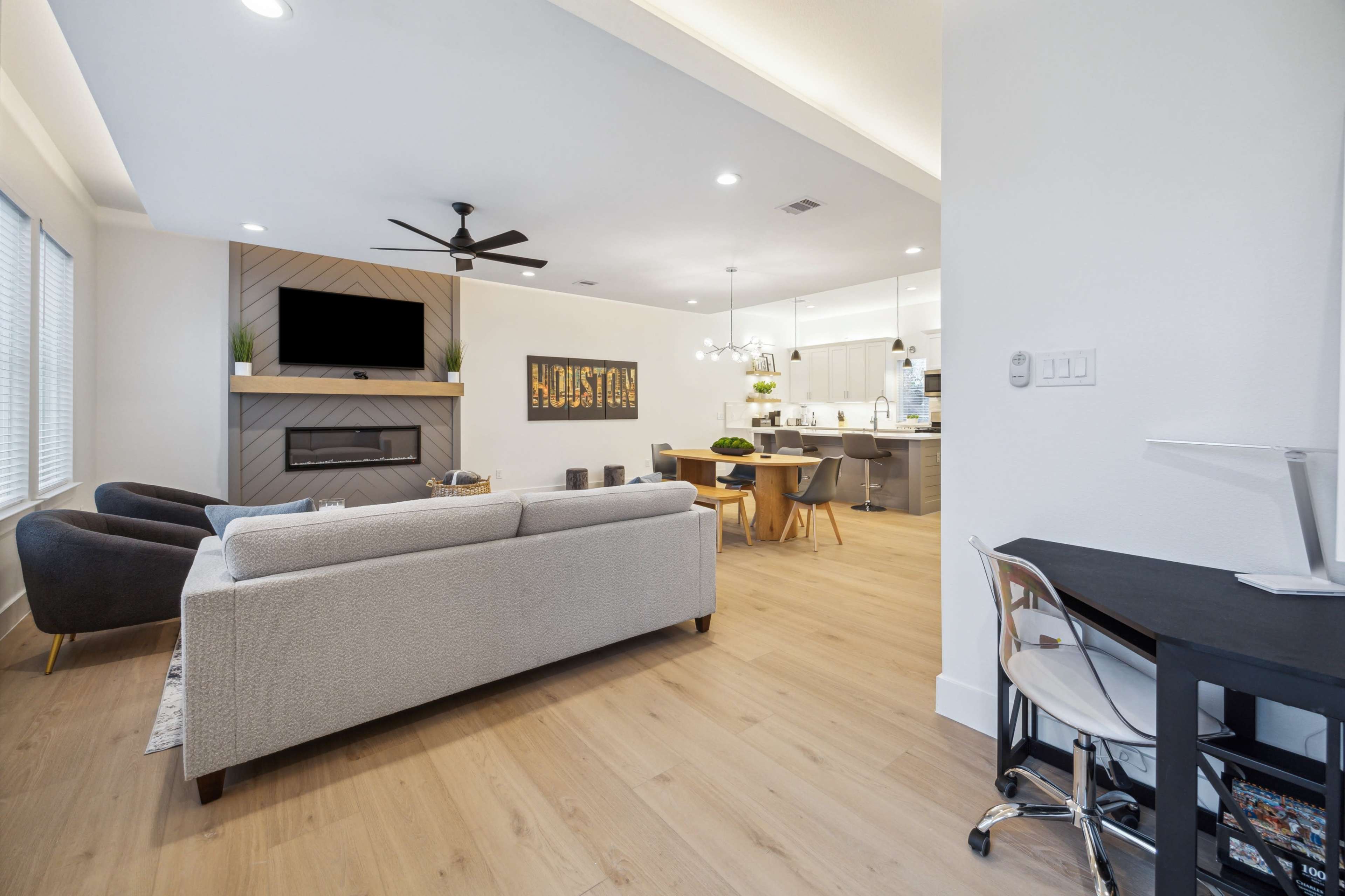A modern living area features a light-colored sectional sofa, a large television mounted above a fireplace, and a dining space with a wooden table and chairs, all set against a backdrop of light hardwood flooring.