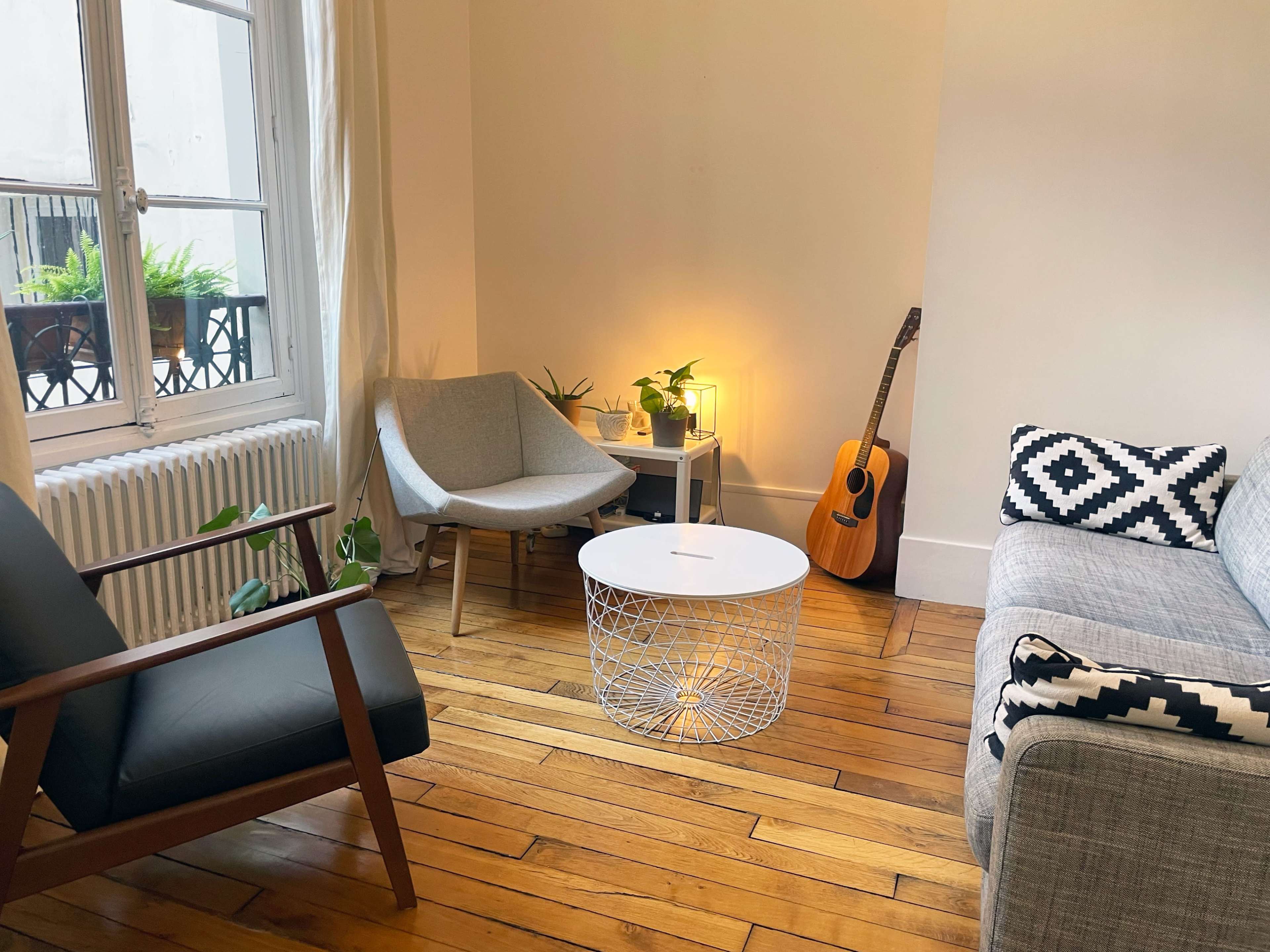 The image shows a cozy living room with a gray sofa, a round white coffee table, an armchair, a guitar, and plants near a window.