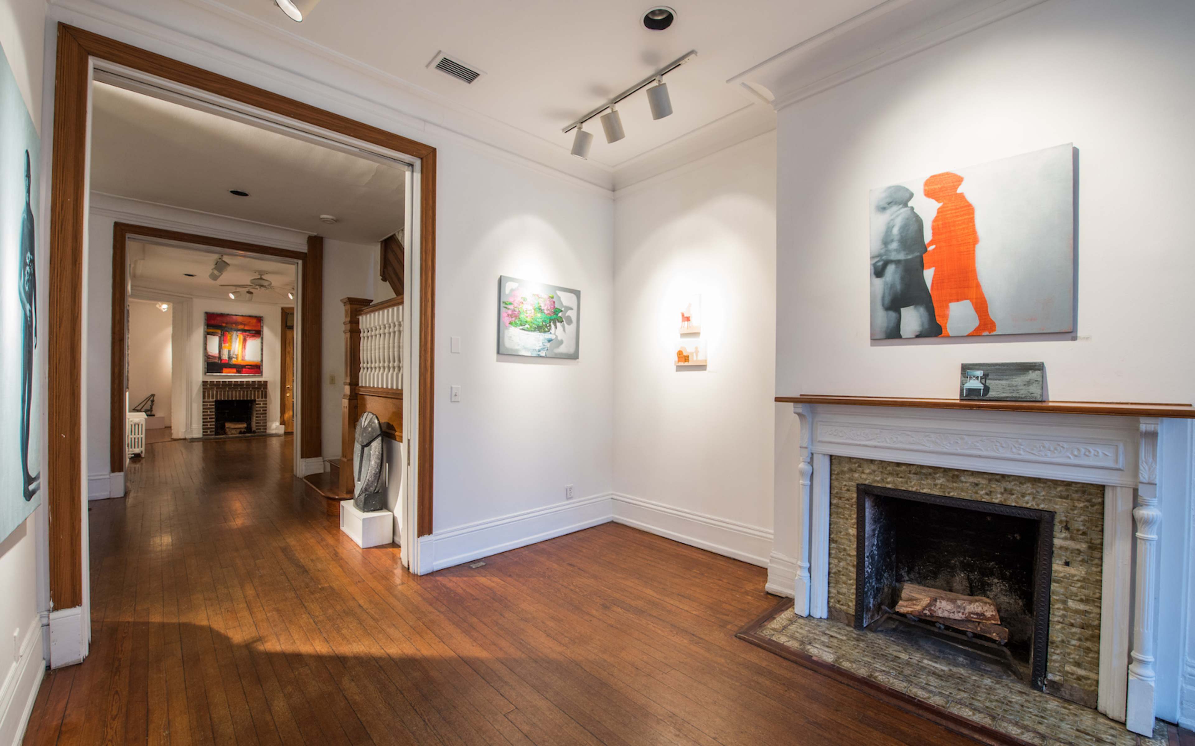 An art gallery with white walls, hardwood floors, and a fireplace, leading to another room featuring more artwork.