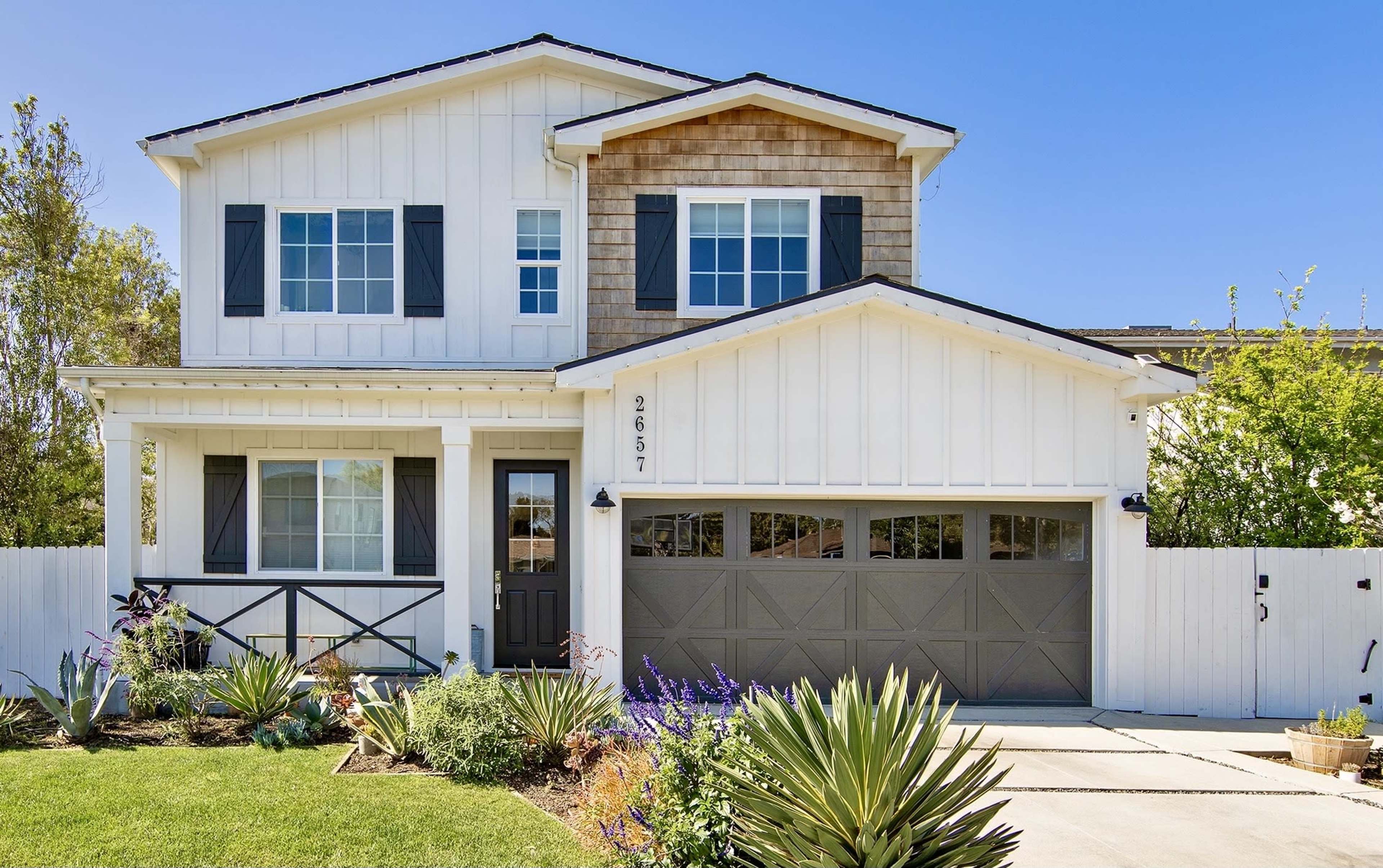 A two-story white house with a brown shingle accent, black shutters, and a driveway leading to a two-car garage.