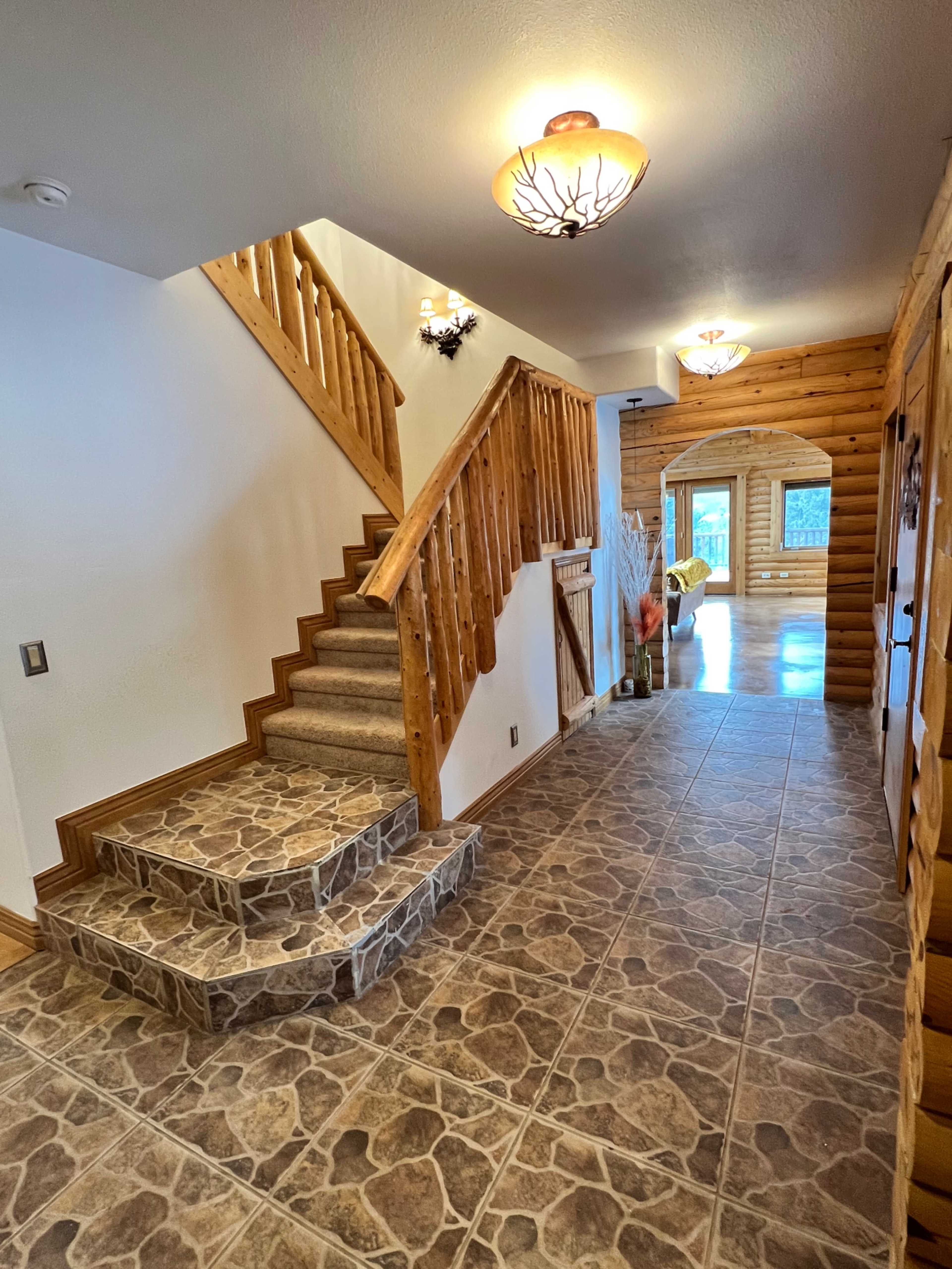 The image shows a spacious hallway featuring a staircase, stone tile flooring, and wooden accents.