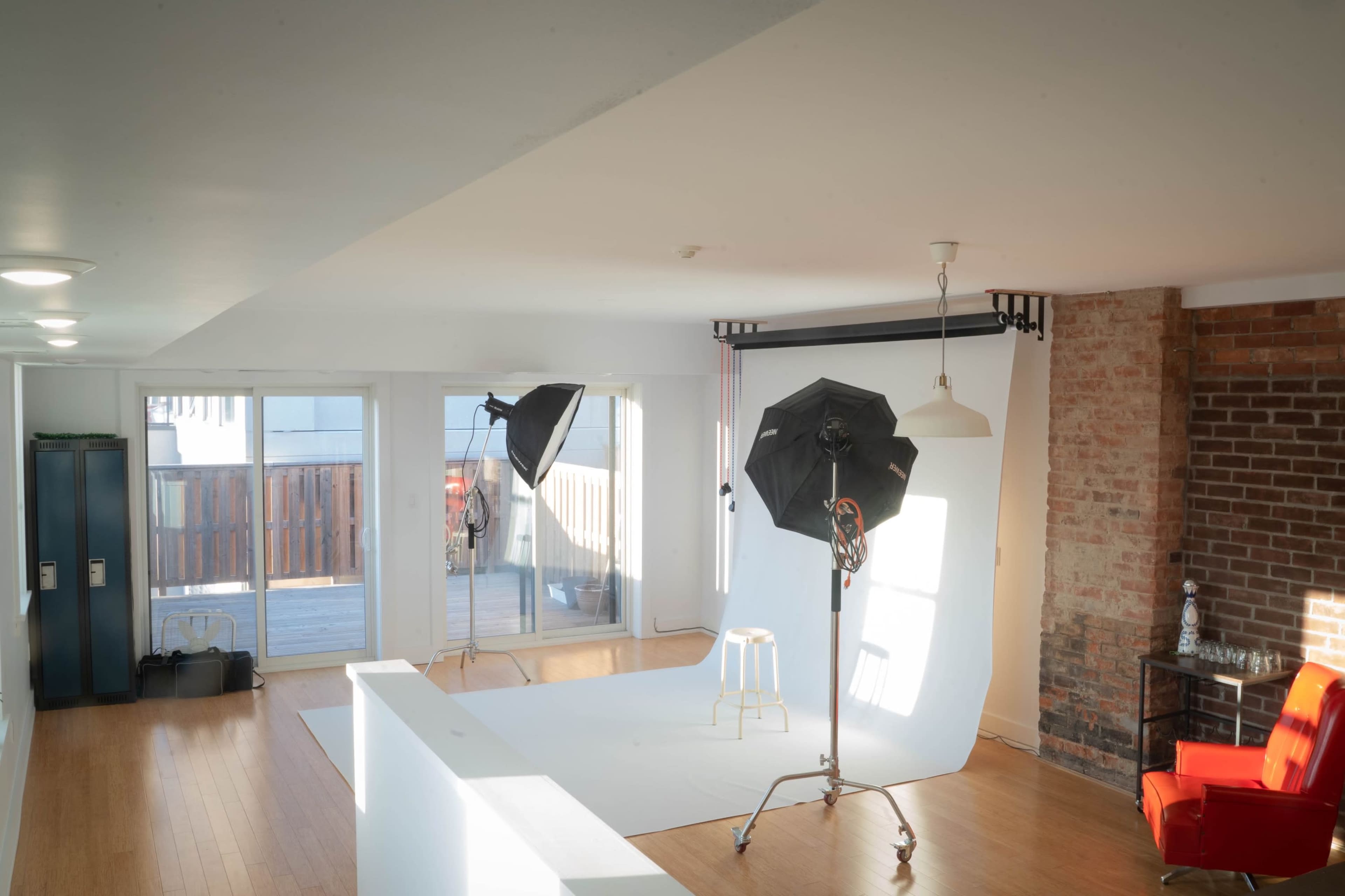 A well-lit photography studio featuring two softbox lights, a white backdrop, and a red chair beside a brick wall.