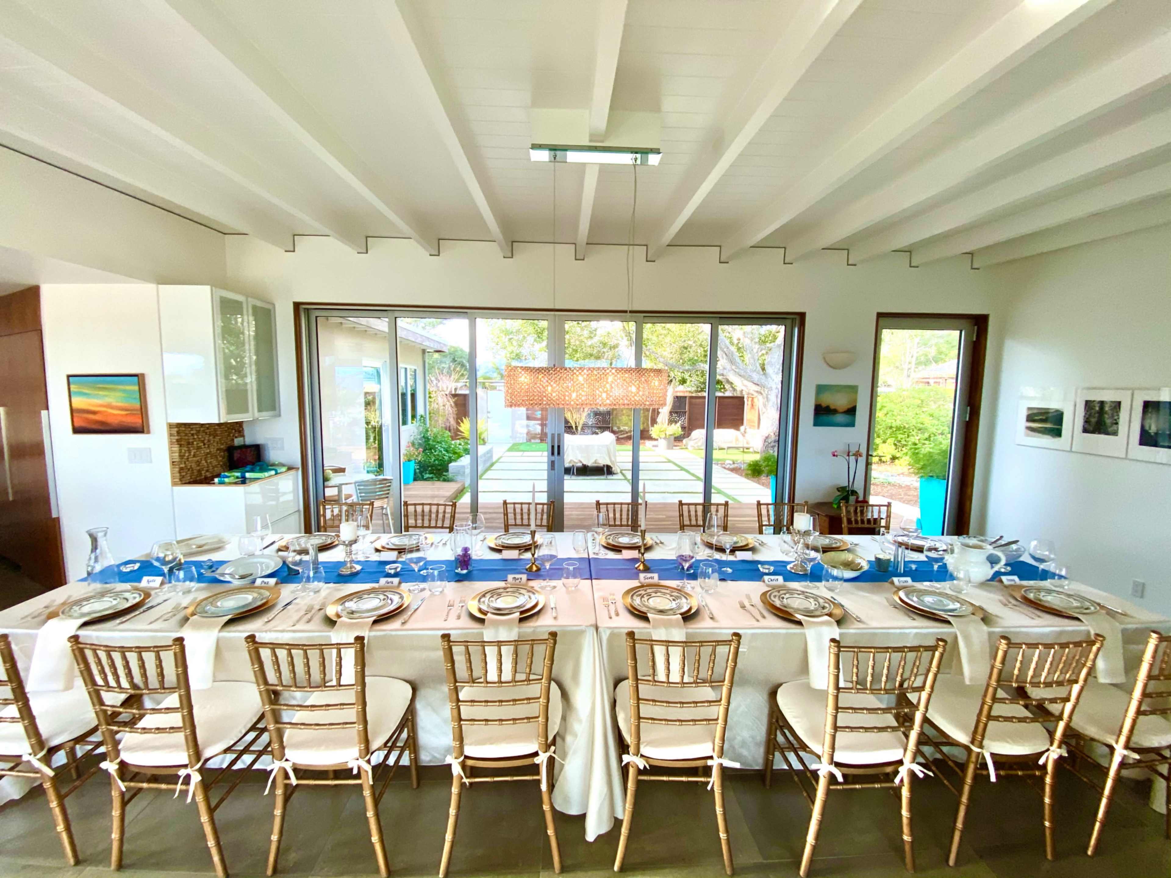 A large dining table is set for a meal in a bright room, with a view of a garden and patio through large glass doors.