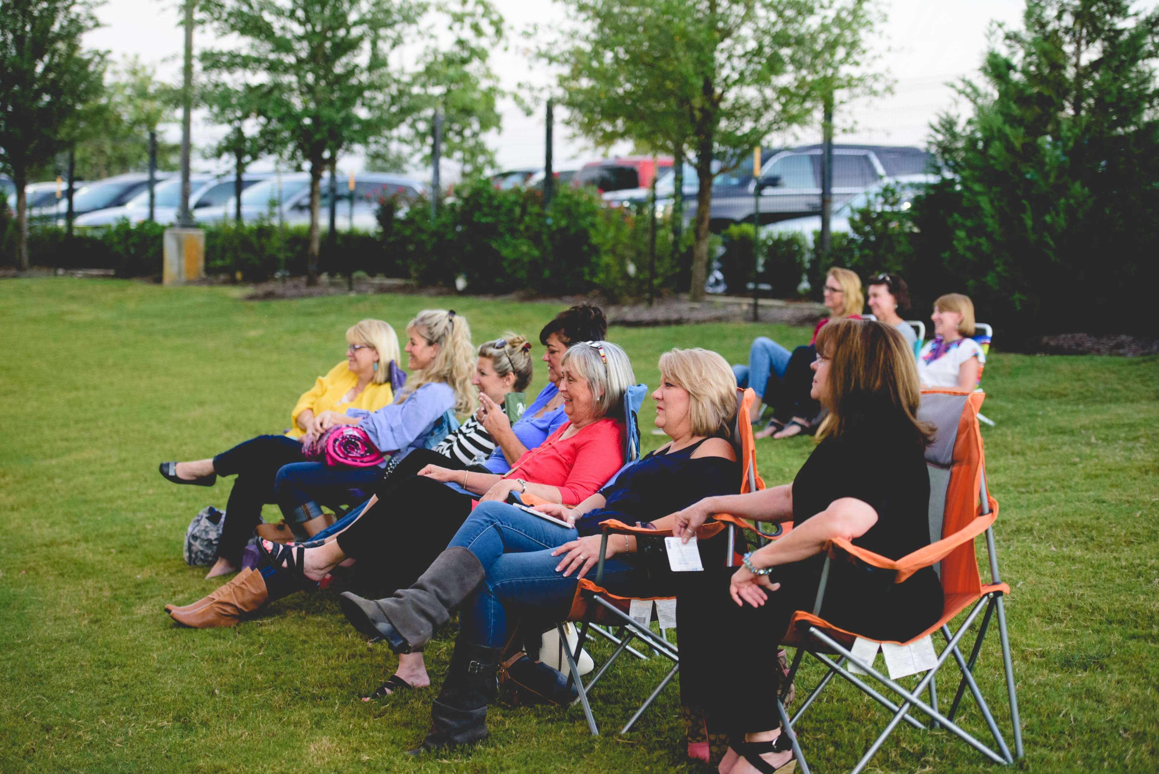 A group of people sit in folding chairs on a grassy area, enjoying a gathering in an outdoor setting.