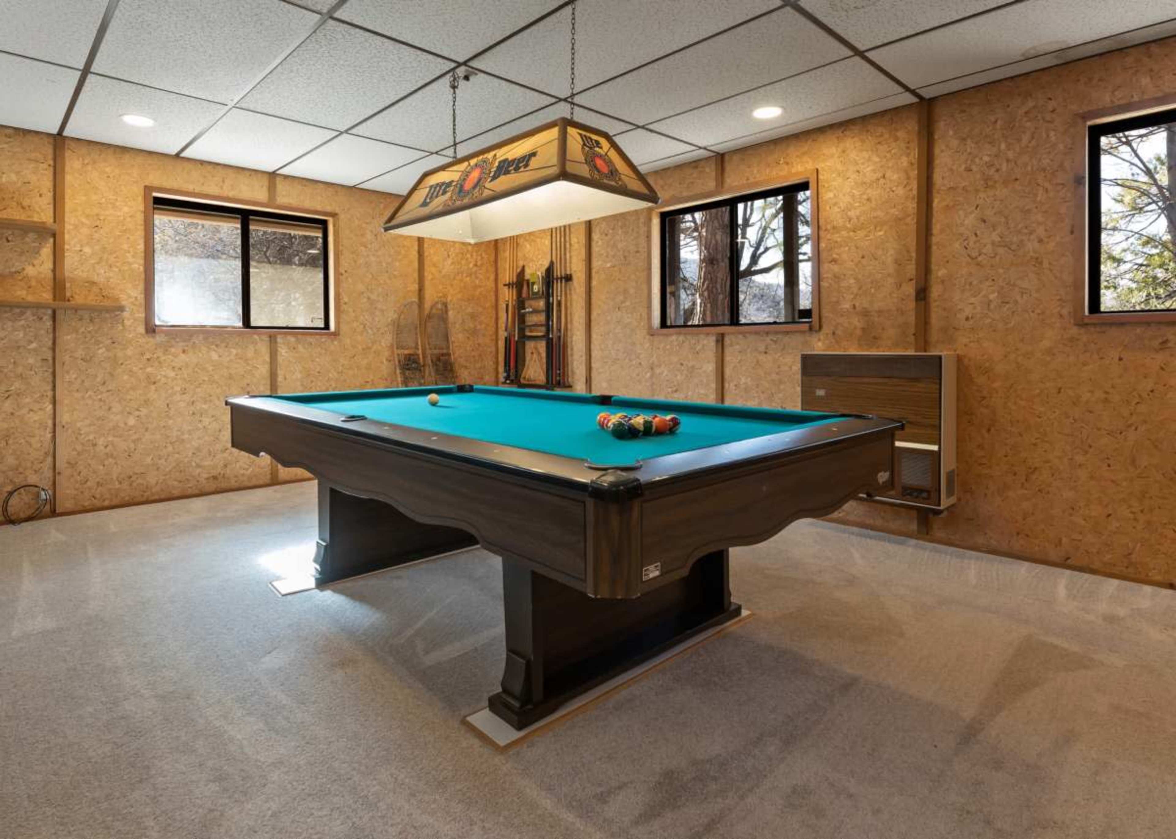 A billiard table with a green felt surface is positioned in a room lined with wood paneling and illuminated by a hanging light fixture.