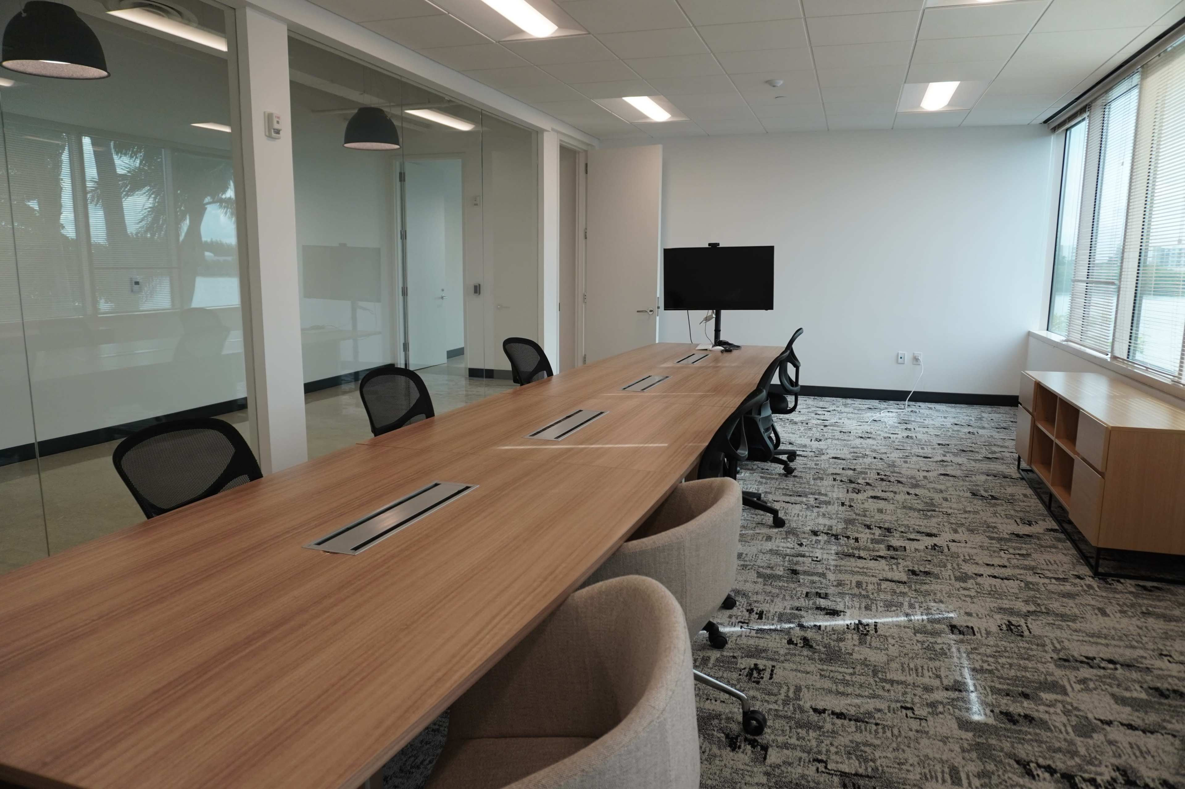 The image shows a modern conference room featuring a long wooden table with several ergonomic chairs, a large screen at one end, and windows providing natural light.