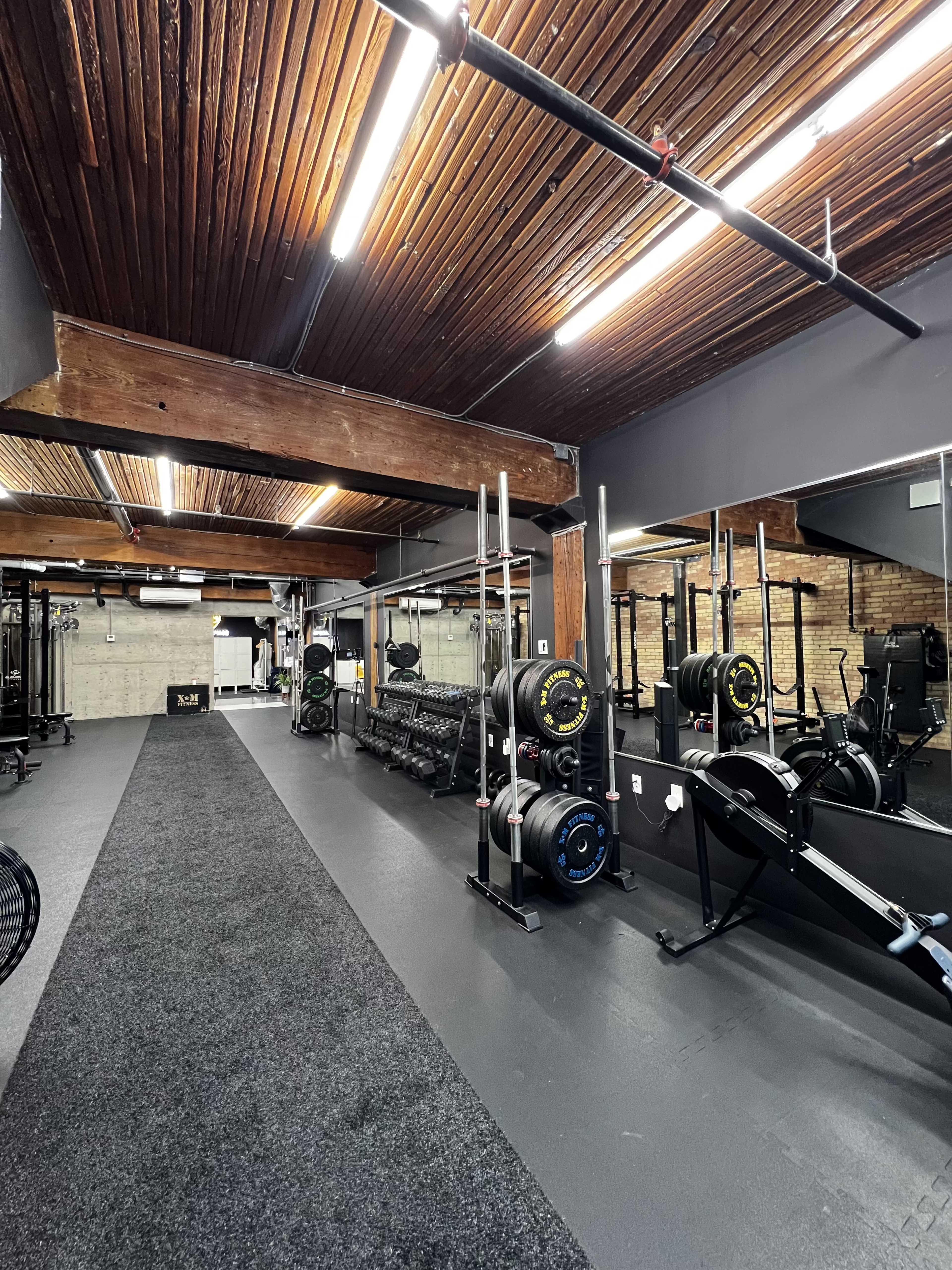 A well-equipped gym interior featuring weightlifting equipment, exercise machines, and a long black carpeted pathway under wooden beams.