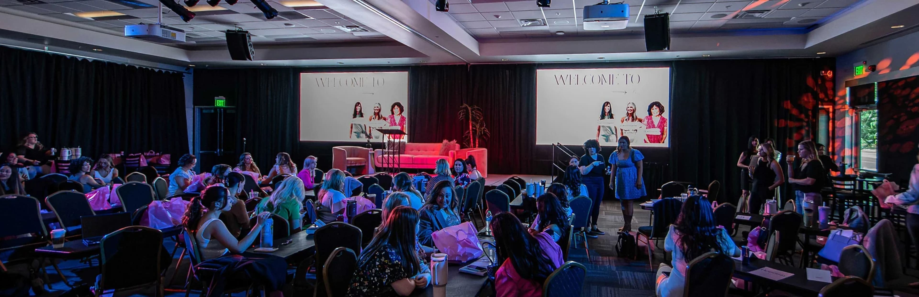 A large conference room filled with participants, seated at tables, facing a stage with a backdrop and a pink couch.