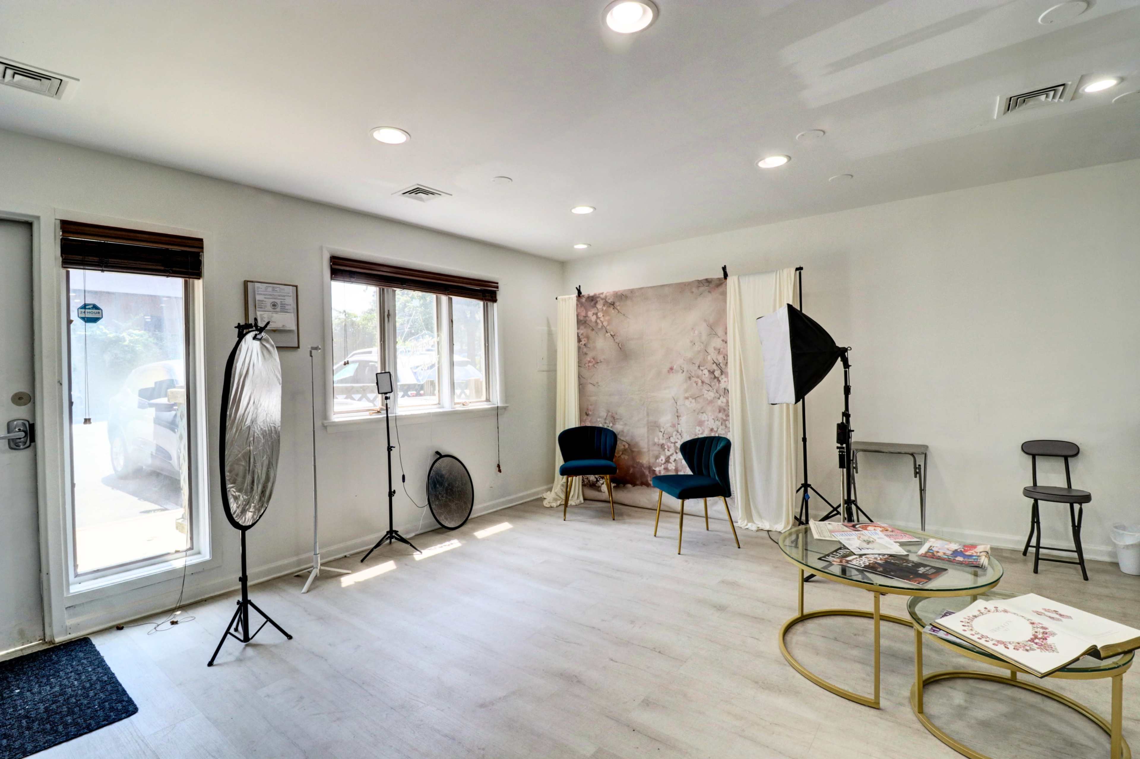 The image shows a bright photography studio with two chairs, a backdrop, a light stand, and various photography equipment arranged in the space.
