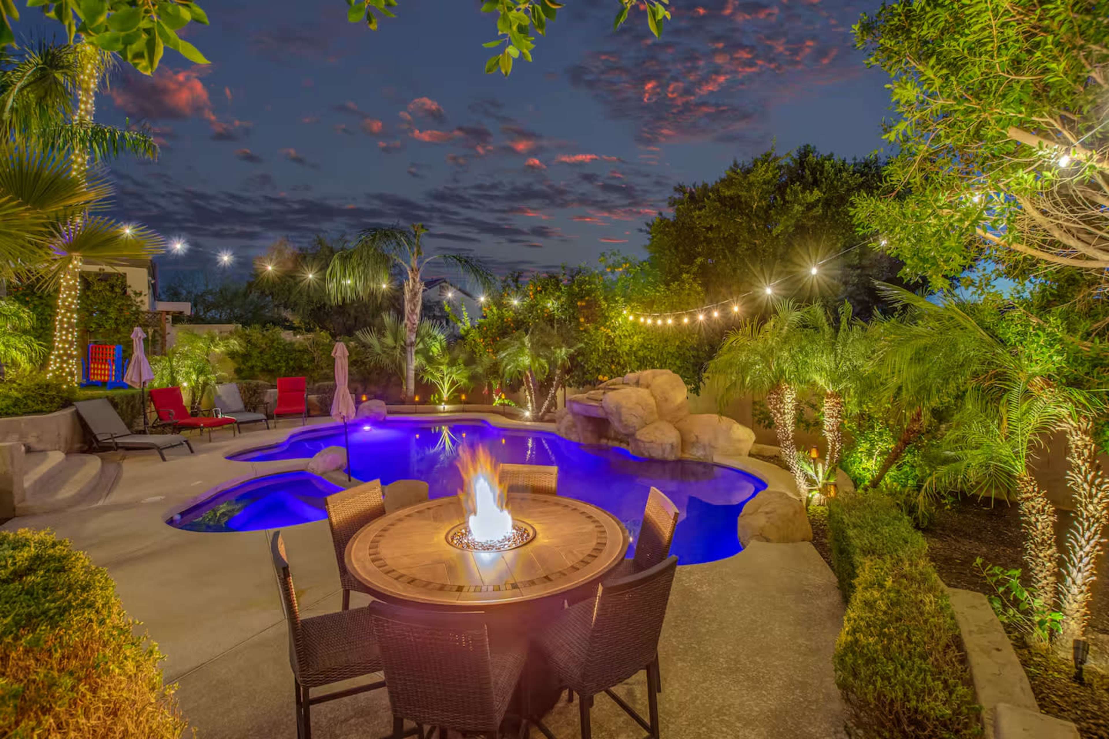 A backyard features a swimming pool surrounded by palm trees, a fire pit at a circular dining table, and string lights overhead.