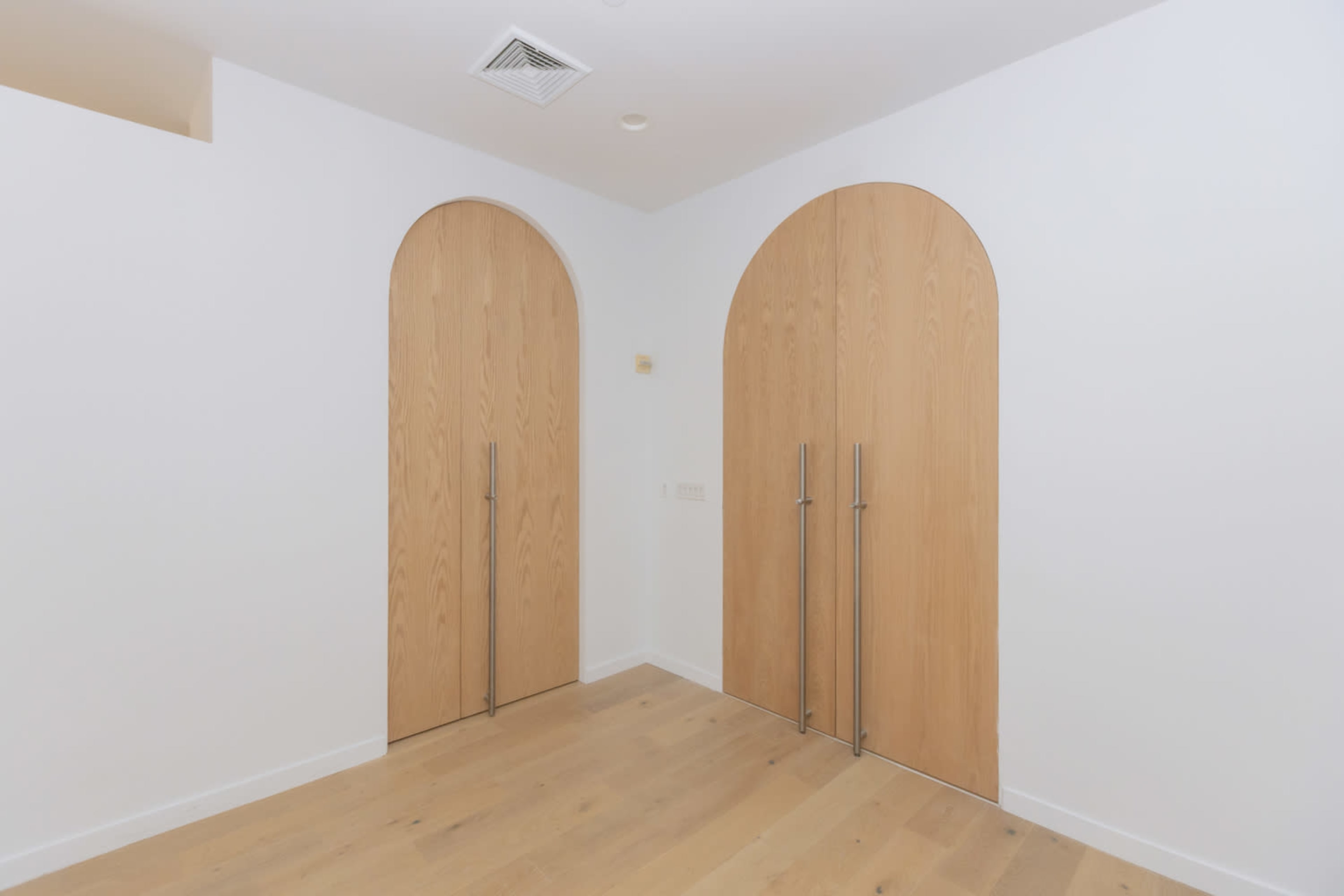 The image shows a minimalistic interior space featuring two wooden doors with arched tops, set against a white wall and wooden floor.