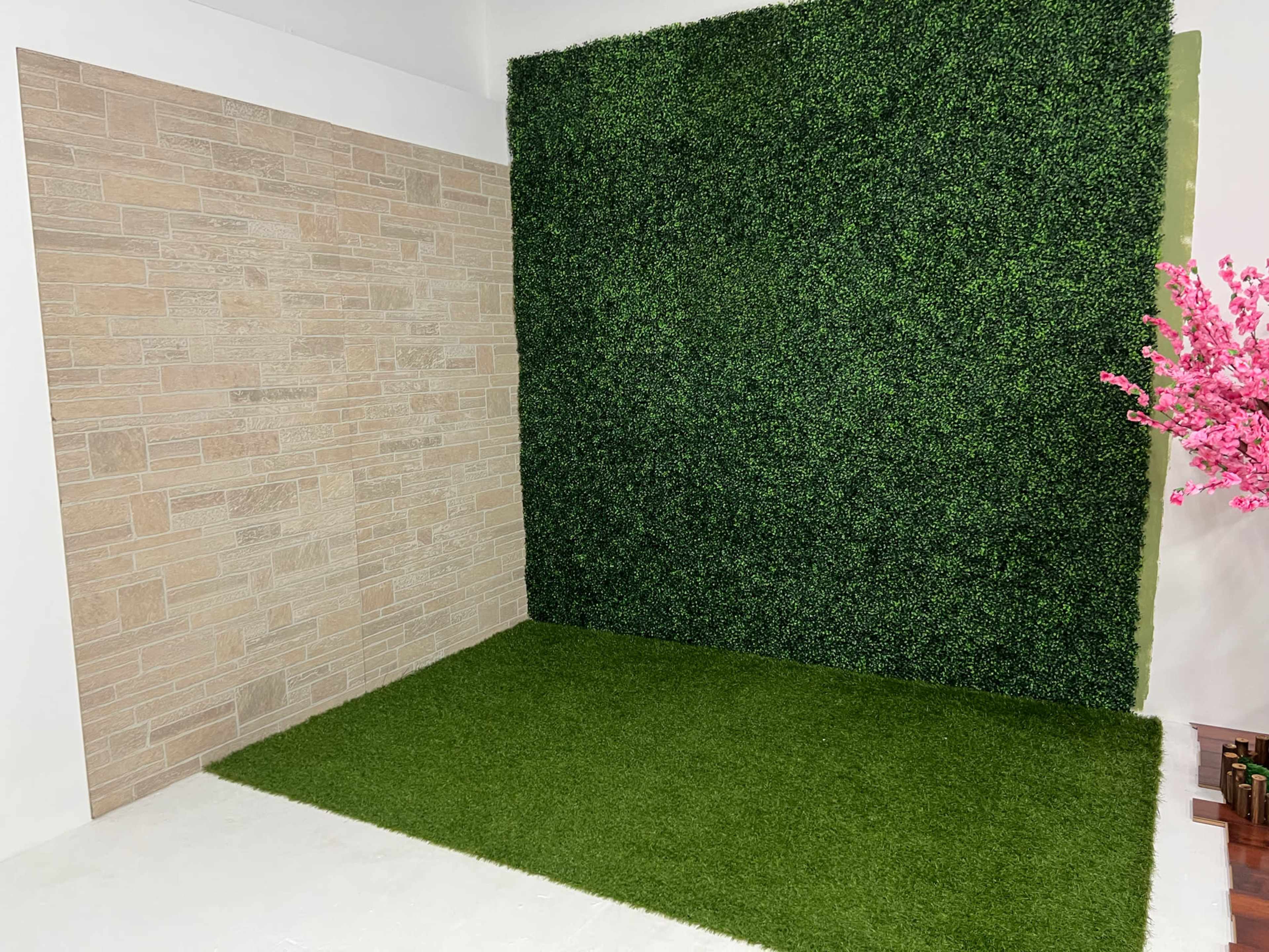 The image shows a staged interior corner featuring a textured stone wall, a green artificial grass area, and a wall covered with faux greenery, along with a pink flowering plant in the foreground.