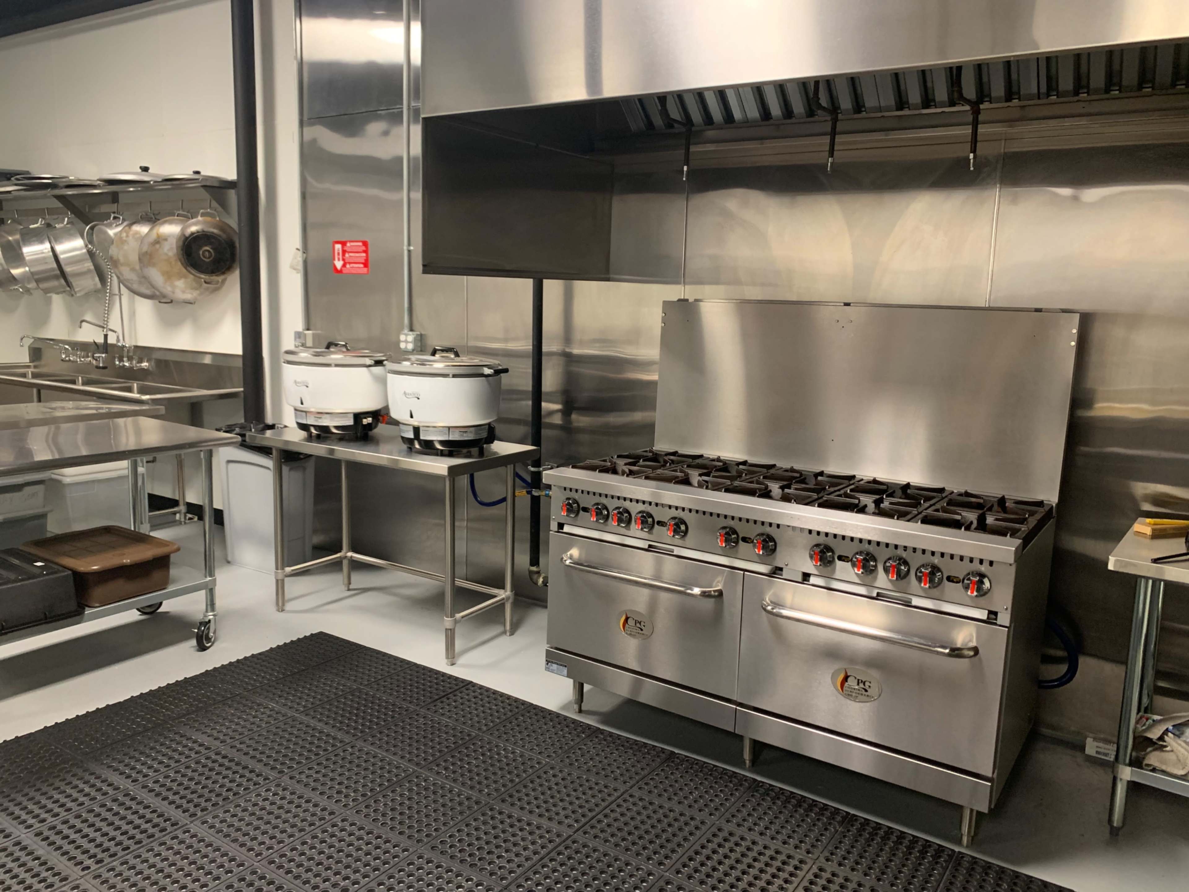 A commercial kitchen with a stainless steel range, a countertop with two food warmers, and a row of hanging pots.