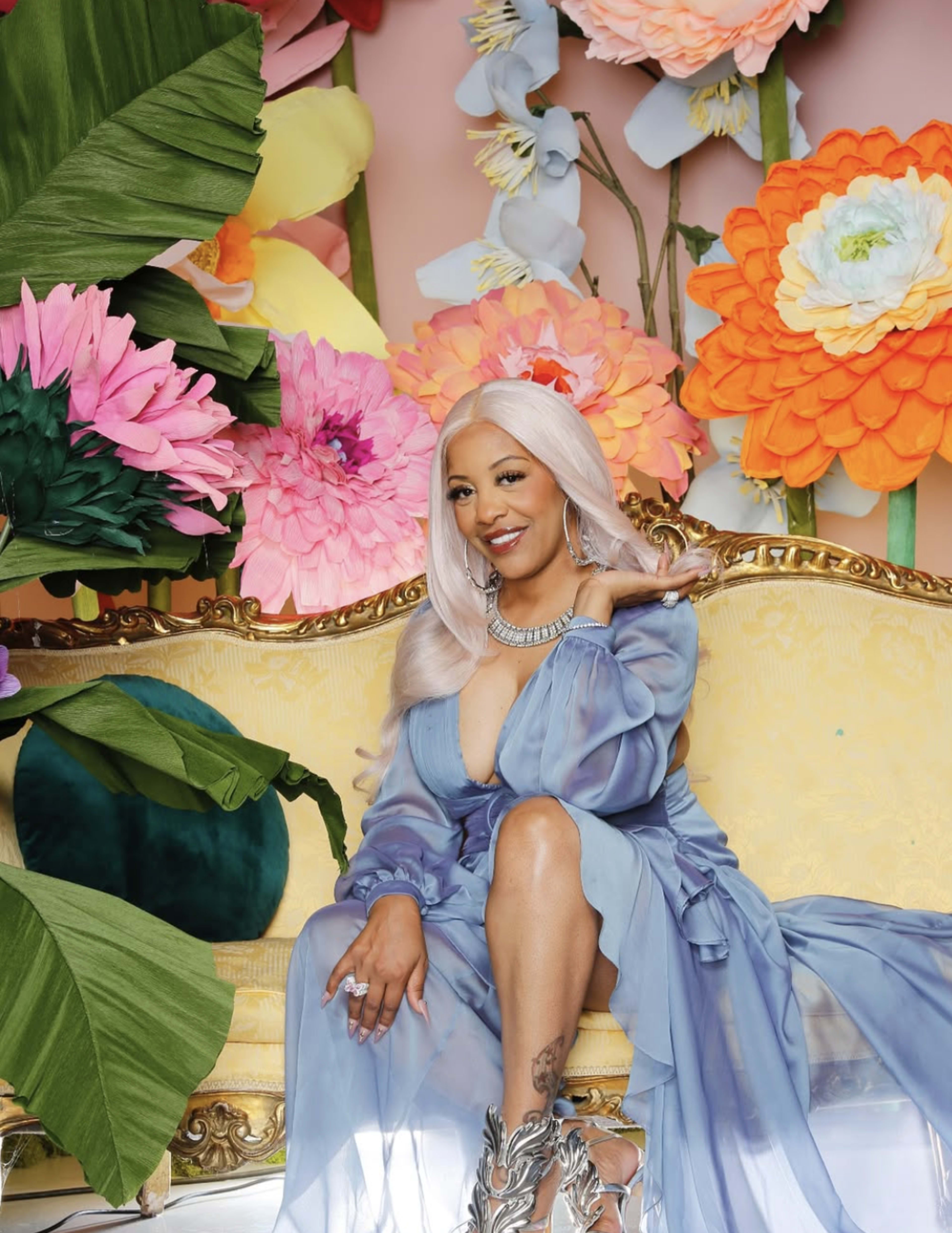 A person with long hair sits on a gold ornate couch surrounded by large, colorful paper flowers.