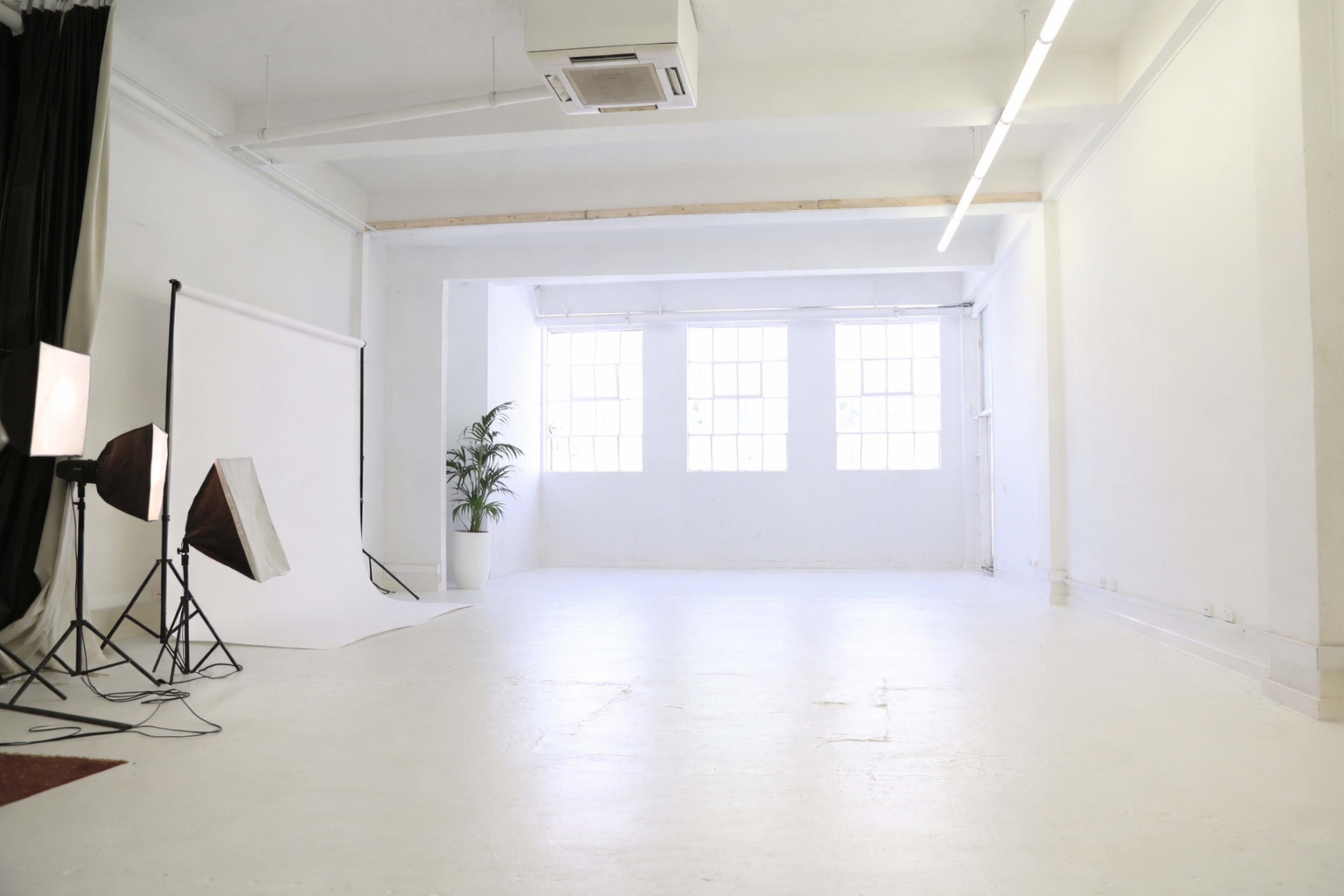 The image shows a spacious, empty studio with white walls, large windows, and minimal lighting equipment set up on one side.