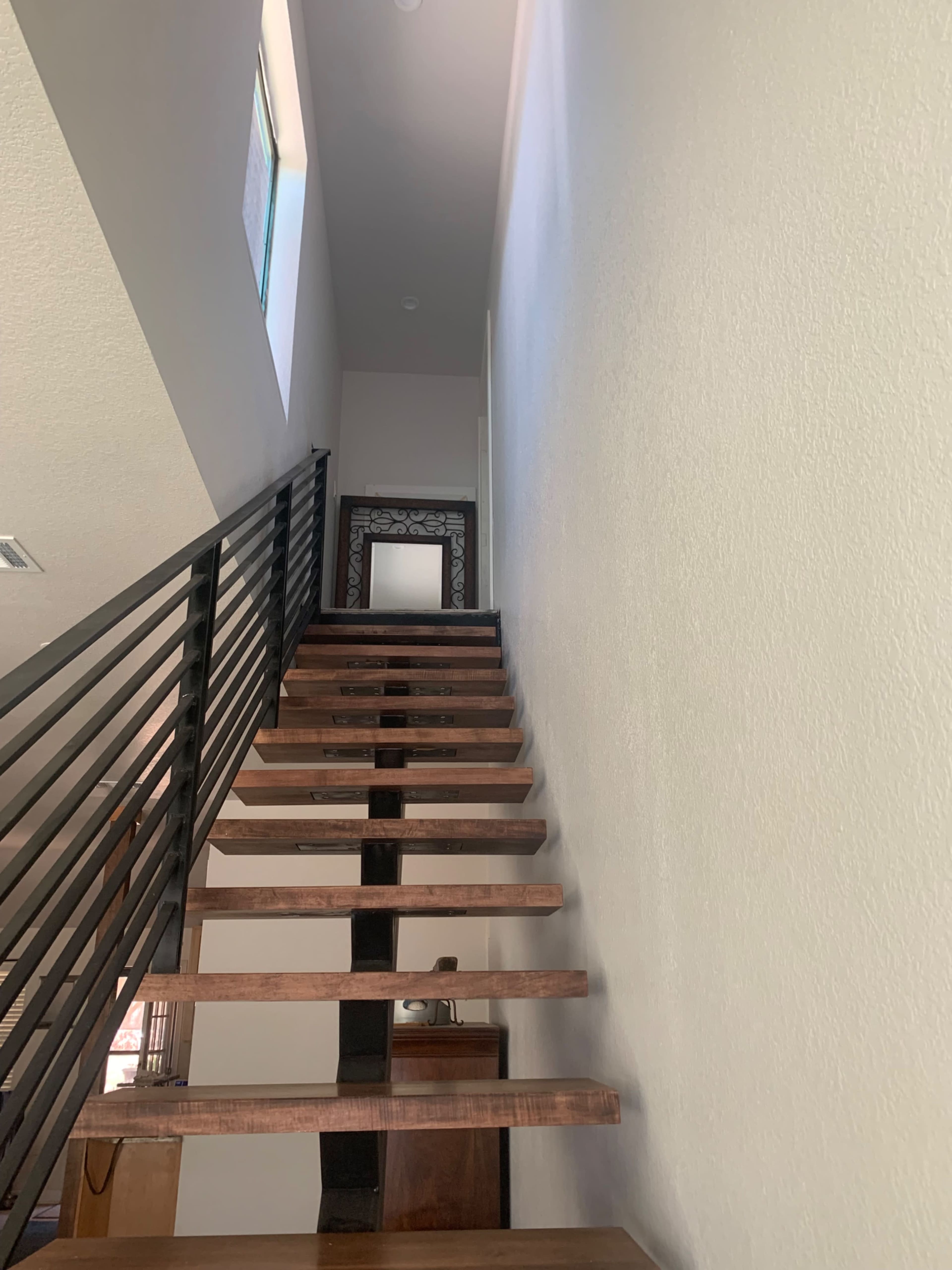 A set of wooden stairs leading up to a landing with a mirror at the top, flanked by a white wall and metal railing.