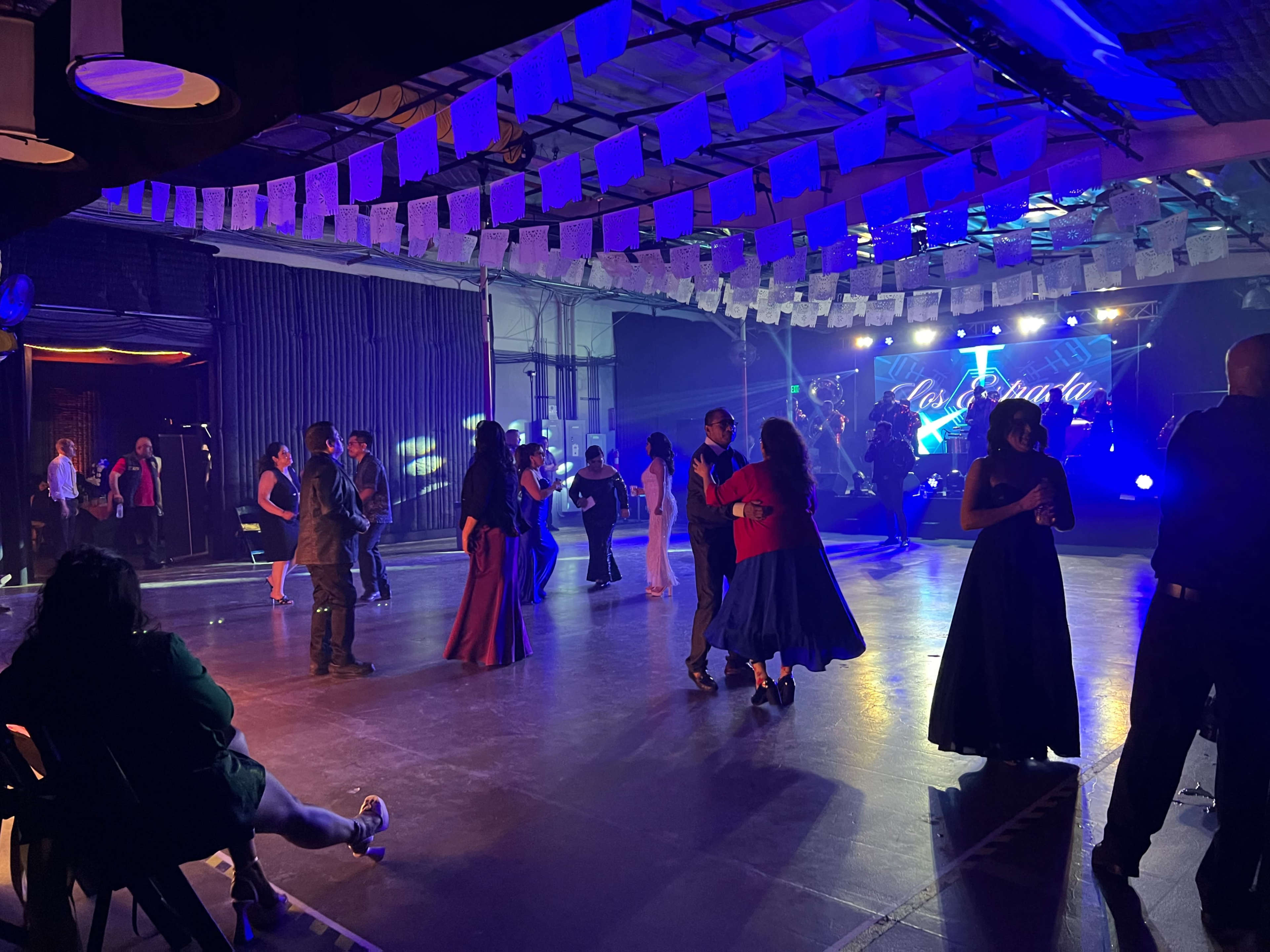 A dance floor is filled with people moving to music under colorful lights, with decorations hanging from the ceiling.