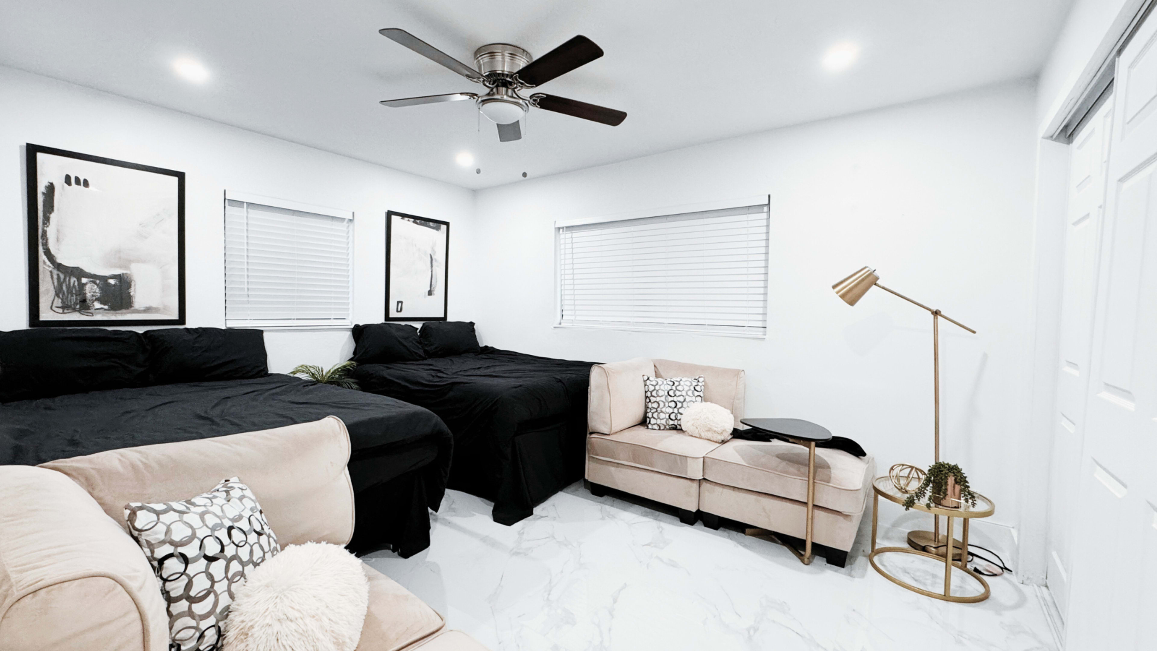 The room features two beds with black linens, a beige sofa with decorative pillows, and a floor lamp, all arranged on marble flooring.