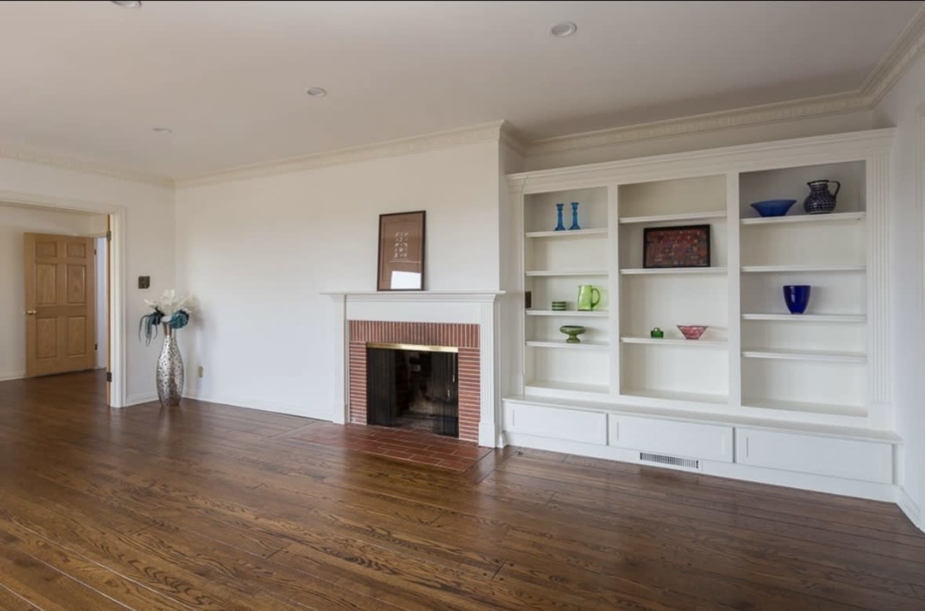A spacious interior room features wooden flooring, a brick fireplace, and built-in shelves displaying various decorative items.